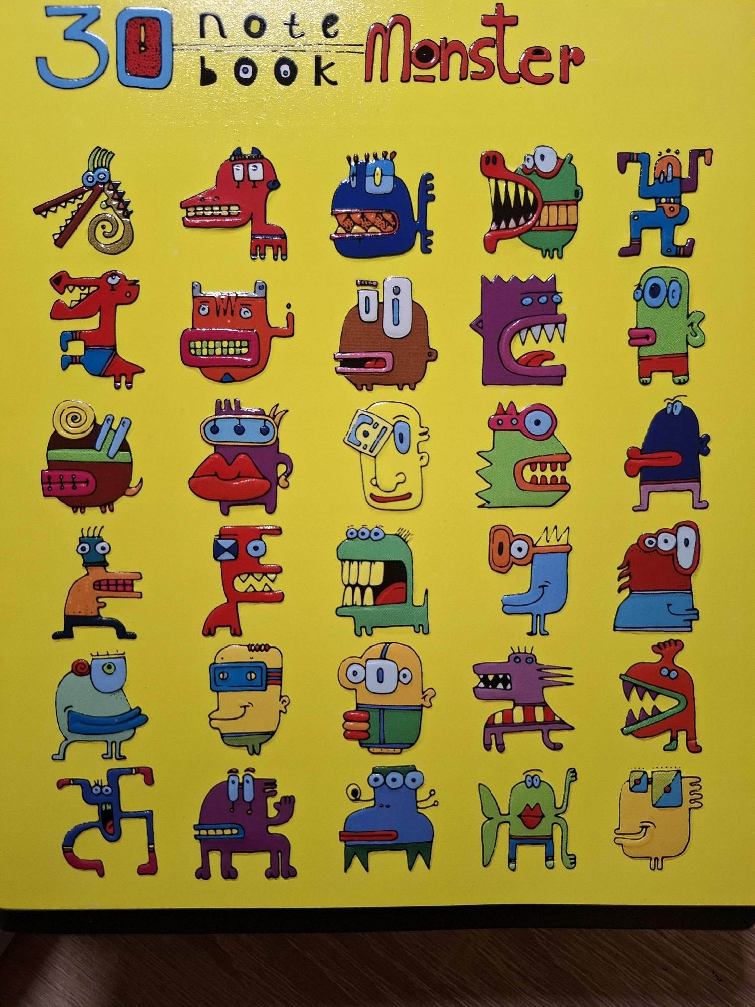 The image is a close-up, overhead shot of a yellow notebook cover. The notebook is titled "30 note Monster" in red and blue lettering. The cover is decorated with a grid of 30 cartoon monster illustrations, each with unique features and colors. The monsters are cartoonish and whimsical, with some having large mouths, multiple eyes, or unusual body shapes. The notebook itself appears to be a physical object, possibly a school or art supply item. There is no indication of the city of Tyumen, Russia, or any specific location within it in the provided image.