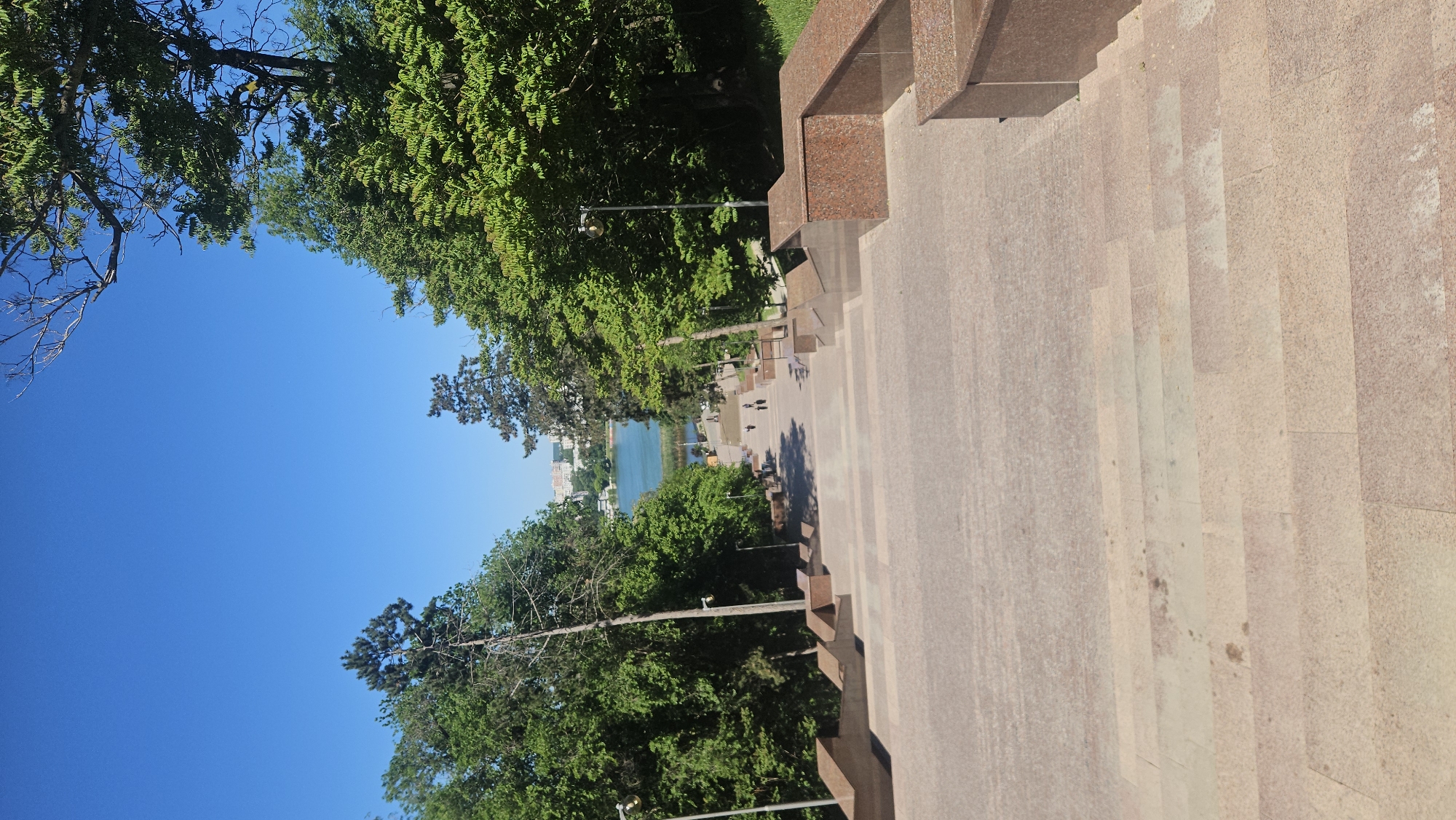 The image depicts a view down a long staircase in Chișinău, Moldova. The staircase is constructed of light-colored stone with darker stone accents on the sides. The view from the top of the staircase reveals a park-like setting with lush trees and a body of water in the distance, presumably a lake or river. The sky is a clear blue. Several people are walking along a path between the stairs and the lake. Street lights are visible in the park area.