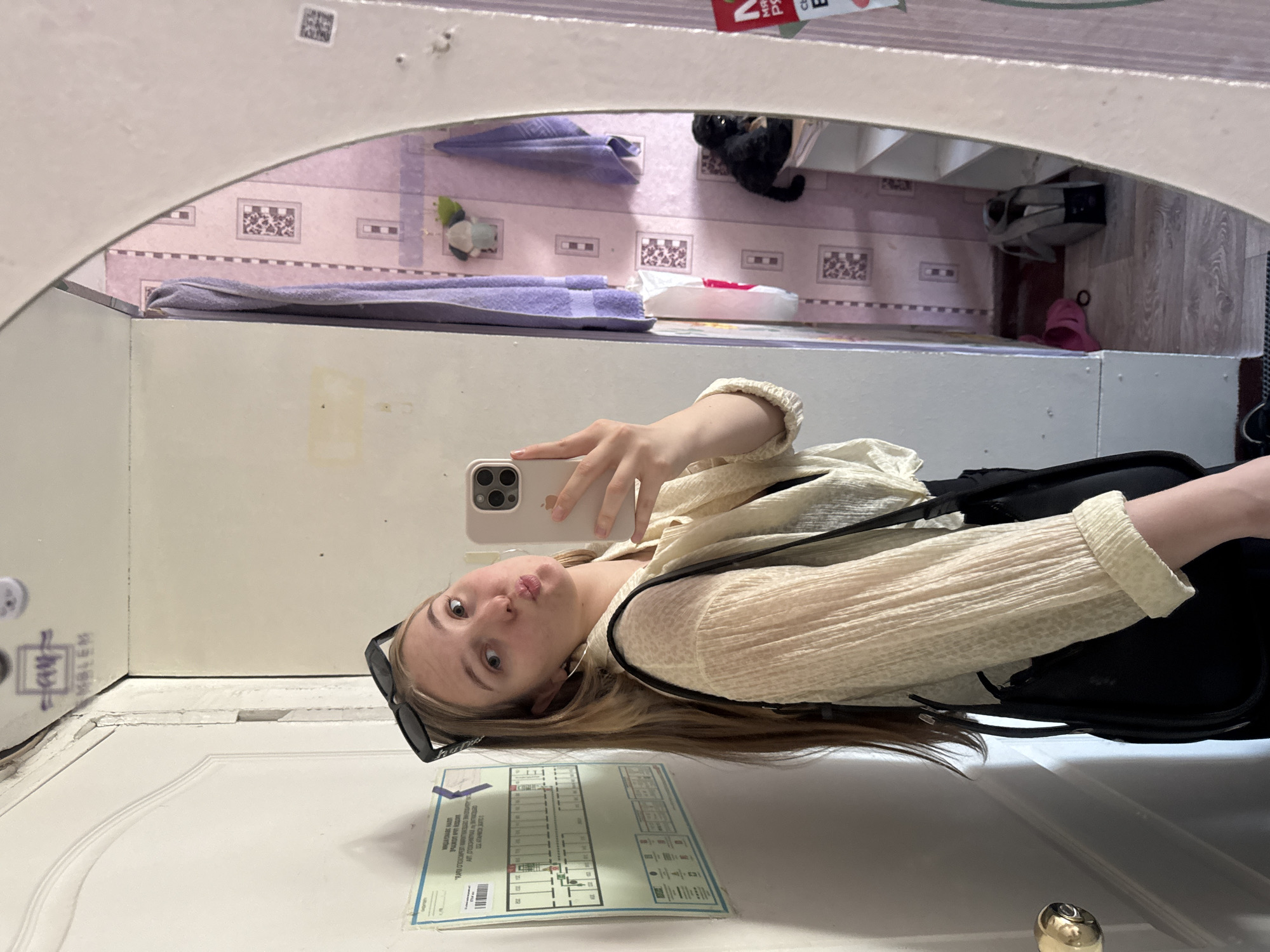 A young woman with long blonde hair is taking a mirror selfie in what appears to be a hallway or bathroom. She is wearing sunglasses on her head, a cream-colored, sheer, crinkled blouse, and a black skirt or pants. A black purse with a long strap is slung across her body. She is puckering her lips as if blowing a kiss.
The mirror shows a reflection of the room behind her. The wall is decorated with a patterned wallpaper, possibly depicting small framed squares or windows, in shades of pink and purple. Two purple towels are hanging on the wall. To the right, a white shelf unit is visible, with a black stuffed animal or toy sitting on top. On the floor, a pair of pink flip-flops and a gray bag are present.
To the left of the woman, a white door with a golden doorknob is visible. On the door, there is a laminated diagram or map, possibly an evacuation plan or building layout, with text in a foreign language, which appears to be Russian. The overall environment suggests a residential or communal living space.
The user specifically requested details about the location in the city of Perm, Russia. However, there are no visual cues in the image that can definitively place the scene in Perm. The presence of Russian text on the diagram is the only potential indicator of the country, but not a specific city. Therefore, it is impossible to confirm the location as Perm, Russia based solely on the provided image.