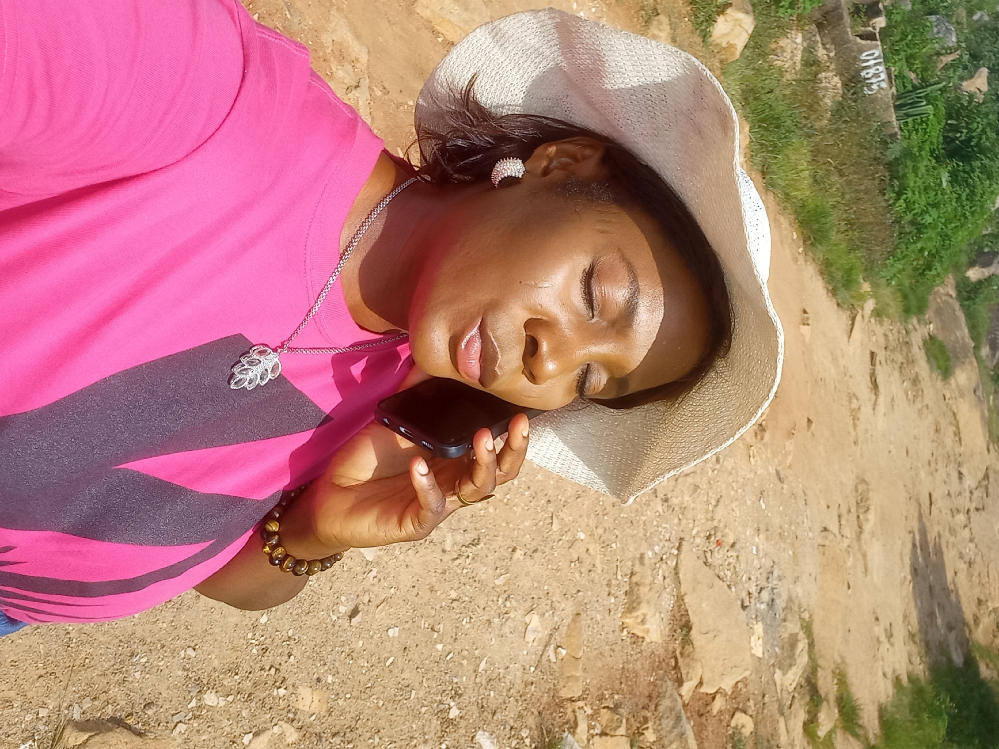 A woman wearing a white sun hat and a pink t-shirt is taking a selfie while holding a black smartphone to her ear. She has her eyes closed and her lips pursed. She is wearing a silver necklace with a leaf-shaped pendant and a bracelet on her right wrist. The background is a dirt path with rocks and sparse vegetation. There is a concrete structure with the number "01875" written on it in the upper left portion of the image.