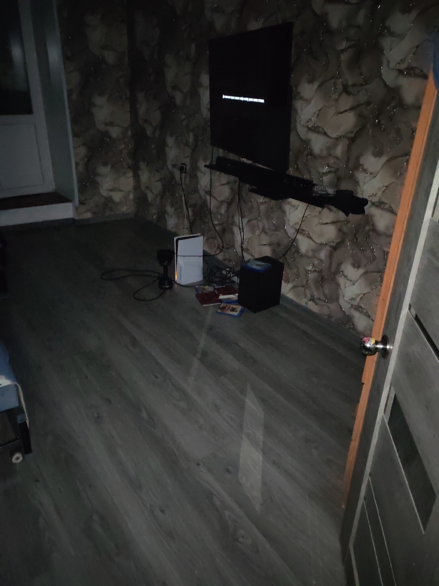 The image depicts the interior of a room in Chulman, Russia. The room is sparsely furnished, with a television mounted on the wall and a gaming console on a shelf below it. A black speaker is placed on the floor next to a stack of what appear to be video games. The walls are covered in a patterned wallpaper, and the floor is made of dark wood laminate. A door is visible on the right side of the image, with a silver doorknob. The lighting is dim, suggesting it is either evening or the room is not well-lit. There are no people or animals visible in the image.