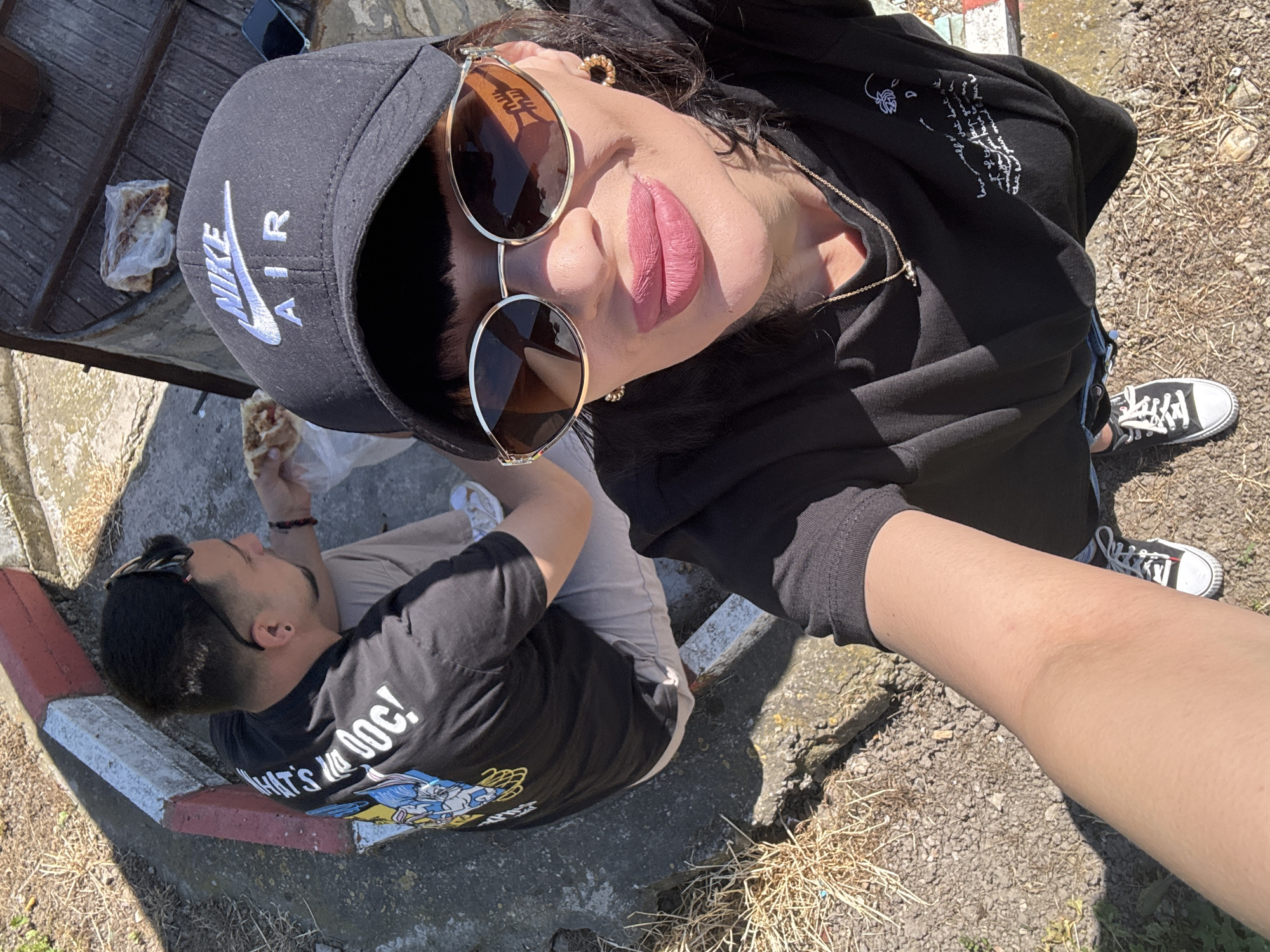 The image, taken from an elevated, near-vertical perspective, depicts two individuals in an outdoor setting in what can be identified as Probuda, Bulgaria.
A person wearing a black Nike Air cap and round sunglasses is positioned towards the top of the frame. They are wearing a black hoodie with white script and a gold necklace, and lipstick, and appear to be holding the camera.
To the left, a second person is seated on a concrete curb with a red and white painted edge. They are wearing a black t-shirt with cartoon characters and the phrase "WHAT'S UP DOC!", light-colored pants, and have sunglasses perched on their head. They are eating food that is wrapped in a plastic bag.
The ground consists of gravel and dry vegetation. In the background, there is a weathered wooden surface with a plastic bag containing remnants of food. The shoes worn by the camera holder appear to be Chuck Taylors. The scene appears to be taken during daylight.