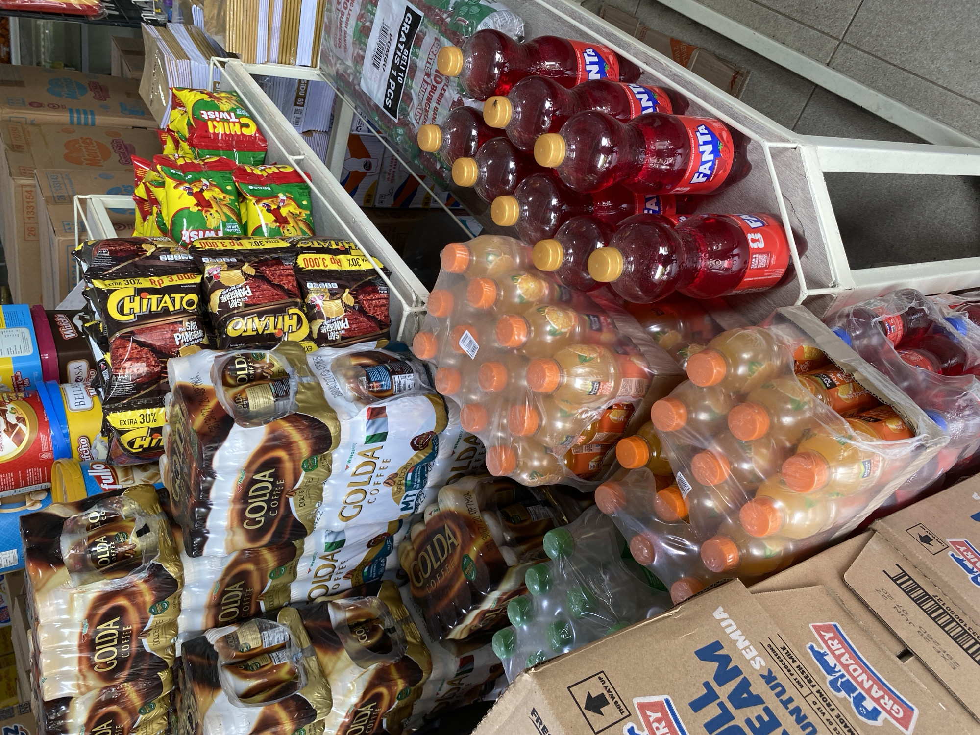 The image depicts a retail store environment in Tanjong, Indonesia, with a focus on beverages and packaged goods. In the foreground, shelves are stocked with various drinks, including several rows of red Fanta bottles, some appearing to be in multipacks. To the left, there are clear plastic bottles containing a light-colored liquid and others with an orange-colored beverage, also packaged in plastic. Several cardboard boxes are visible, including one labeled "FULL CREAM" with "GRAND AIRY" branding, suggesting dairy products. In the background, stacks of packaged coffee products, identified as "GOLDA COFFEE," are prominently displayed. Further back, there are racks of snack items, such as "CHATTO" and "CHIKI TWIST" biscuits. The store appears to have a tiled floor and metal shelving units, typical of a local convenience store or minimarket. No people are visible in the scene.