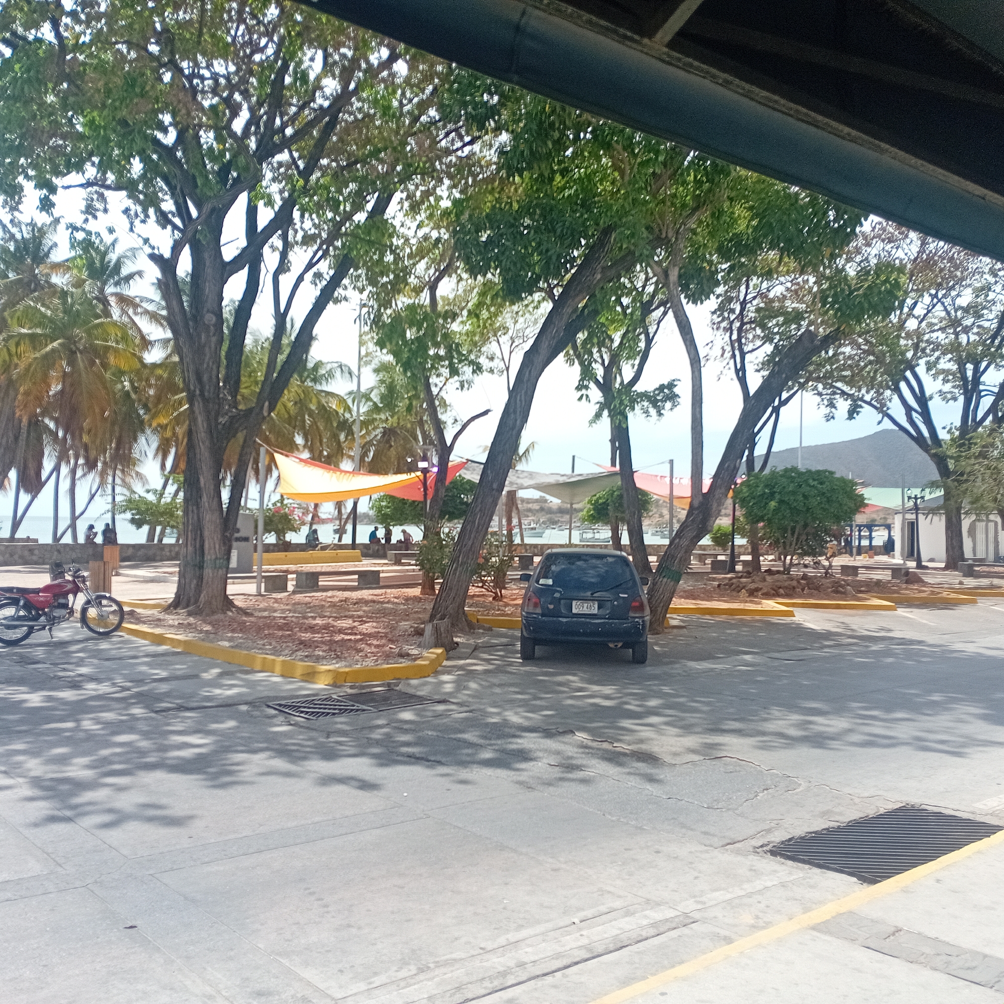 The image depicts a scene on a sunny day in what appears to be a public area. A dark-colored car is parked between trees in a parking area marked with yellow curbs. A red motorcycle is parked nearby. In the background, there is a waterfront area with palm trees, canopies, benches, and people. A mountain is visible in the distance. The foreground features a street surface with shadows from the trees. The location is Unknown, Unknown.
