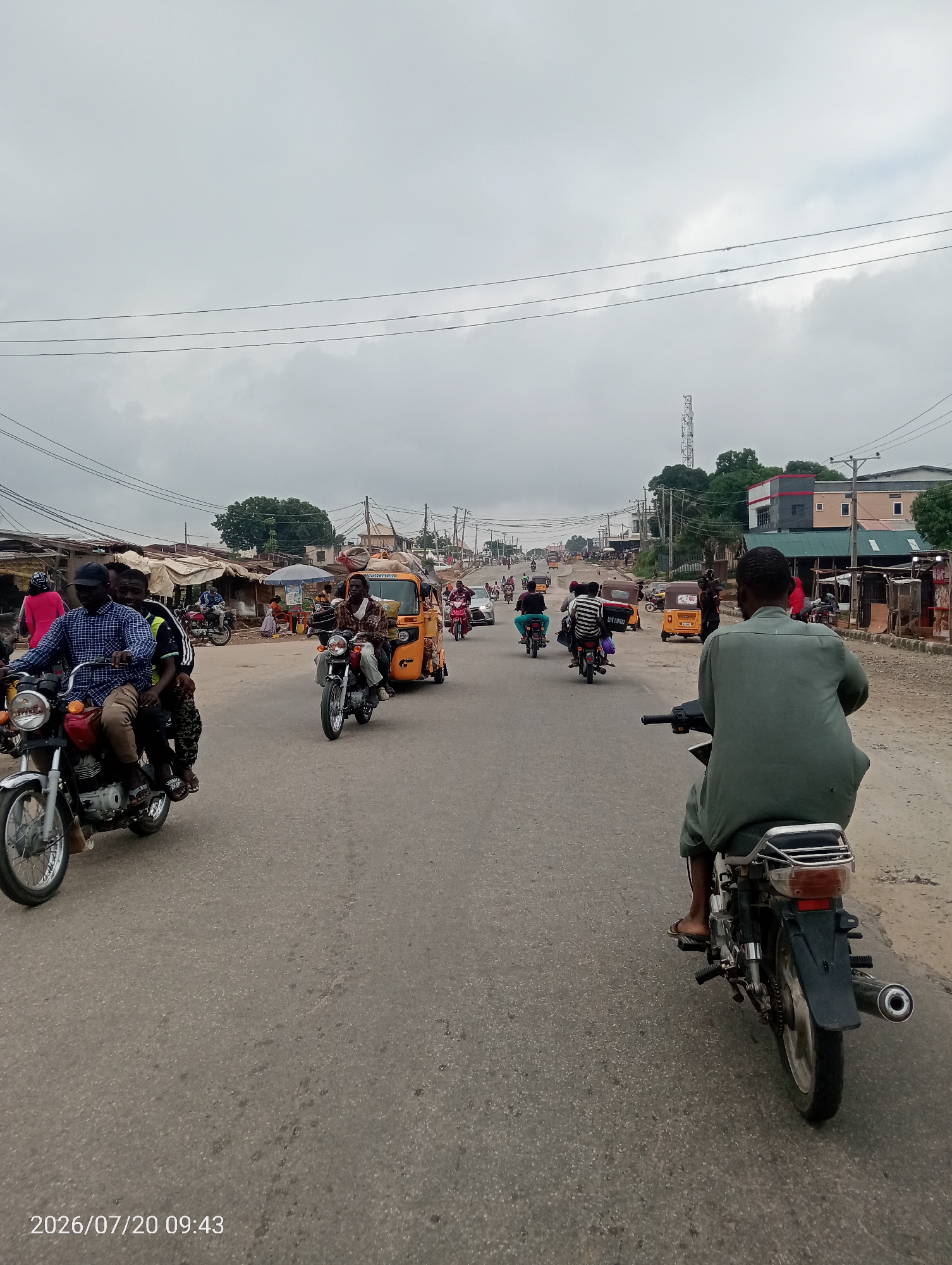 The image depicts a busy street scene in Suleja, Nigeria, on July 20, 2026, at 09:43. The sky is overcast, suggesting a cloudy day. The road is paved and appears well-trafficked by various vehicles. In the foreground, a person is riding a motorcycle, viewed from behind. To the left, another motorcycle carries three people, with the rider in a blue and white checkered shirt and a black cap. Further down the road, an orange auto-rickshaw, or "keke," is visible, loaded with goods. Several other motorcycles and auto-rickshaws are present on the road, indicating active local transportation. The sides of the road are lined with various structures, including market stalls, shops, and buildings, suggesting a commercial and residential area. Power lines are visible overhead, crisscrossing the sky. The overall impression is of a typical day in a bustling Nigerian city.