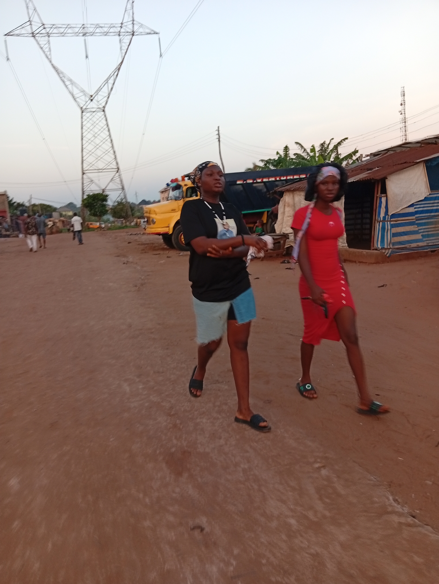 Two young women walk on a reddish-brown dirt road in Ogidi, Nigeria. The woman on the left wears a black t-shirt, a light blue denim skirt, and sandals. She has a headscarf and a necklace. The woman on the right wears a red dress with buttons, a headscarf, and sandals. In the background, there is a large yellow truck with equipment on top, power lines on a metal tower, and simple buildings. Other people are walking on the road in the distance.