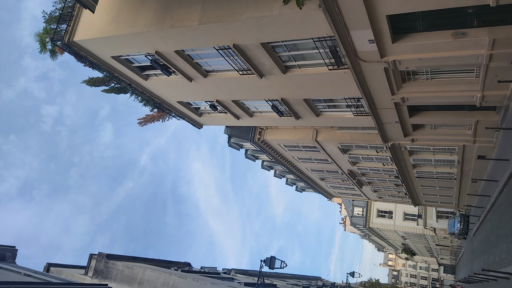 The image depicts a street scene in Paris, France, likely in a residential area characterized by Haussmannian architecture. The perspective is low, looking up along a narrow street lined with tall buildings on either side. The sky above is a pale blue with faint white clouds and a subtle contrail.
On the left, the facade of a building is partially visible, showing a dark, traditional street lamp attached to its side. The building is clad in a light gray material.
The right side of the street is dominated by a series of cream-colored buildings with steeply pitched mansard roofs, typical of Parisian construction. These buildings have multiple stories, each featuring numerous windows with white shutters. Some windows have small black metal railings or flower boxes. The building in the foreground on the right is taller and wider than those to its left. It has a smooth facade and a number "9" is visible above one of its doorways. There is greenery and trees on the rooftop of the tallest building.
The street itself is paved with cobblestones. A silver car is parked on the left side of the street. Several black metal bollards are lined up along the sidewalk on both sides of the street. A couple of figures are faintly visible walking in the distance on the left sidewalk. The overall impression is a quiet, early morning or late afternoon atmosphere in a classic Parisian neighborhood.