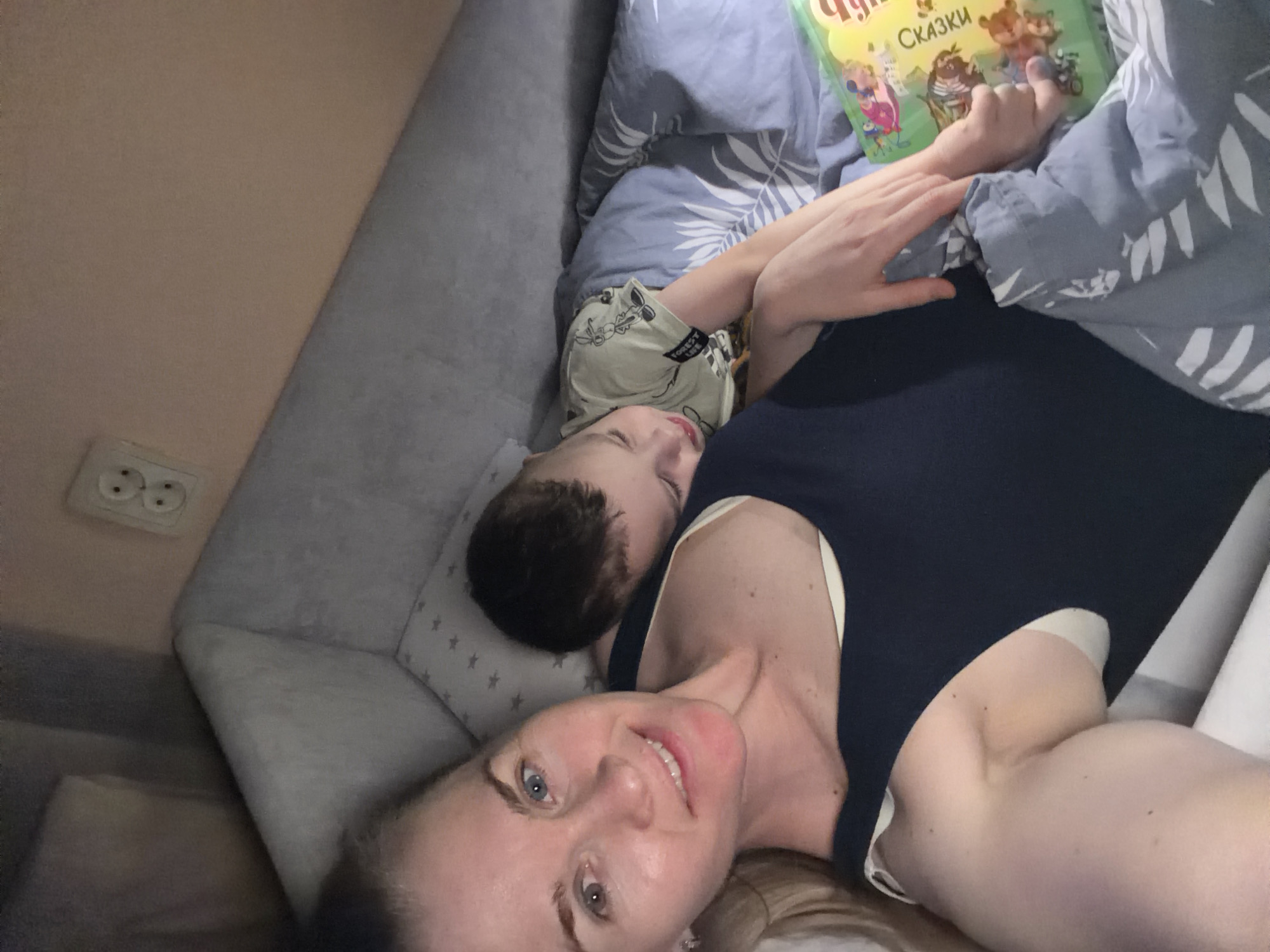 A woman and a young child are lying on a sofa. The woman is taking a selfie, looking at the camera with a smile. The child is lying on their side, with their head resting on a pillow. They appear to be asleep or drowsy. The child is wearing a light-colored t-shirt with a cartoon print.
In the background, a book is held by a person's hand, partially obscuring the child. The book has a colorful cover with cartoon characters and text that appears to be in Russian. The word "СКАЗКИ" (Skazki, meaning "Fairy Tales") is visible on the book cover.
The setting is indoors, likely a living room. A wall with a power outlet is visible to the left of the sofa. The lighting suggests it could be evening or nighttime. The mood is intimate and peaceful, indicative of a quiet moment between a mother and child. The woman's expression is warm and loving. The image is taken from a high-angle perspective.