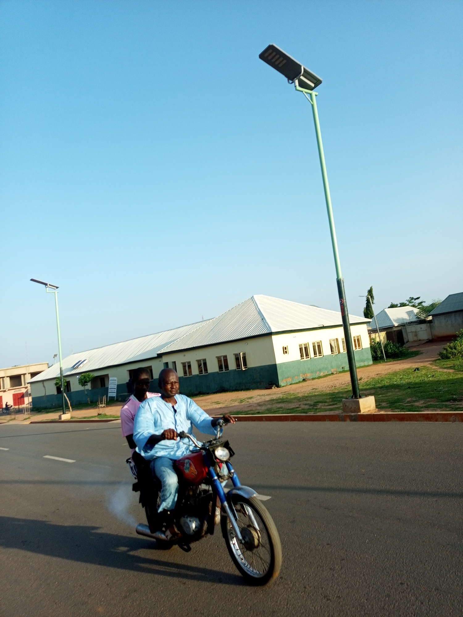 Two men are riding a red motorcycle on an asphalt road. The man in the front is wearing a light blue patterned shirt and blue jeans. The man behind him is wearing a pink shirt. The motorcycle is emitting smoke from its exhaust. In the background, there is a light-colored building with a white roof and green trim. A tall, green lamppost with a solar panel is visible to the right of the building. The sky is clear and blue. The scene appears to be in Pantame, Nigeria, based on the provided context.