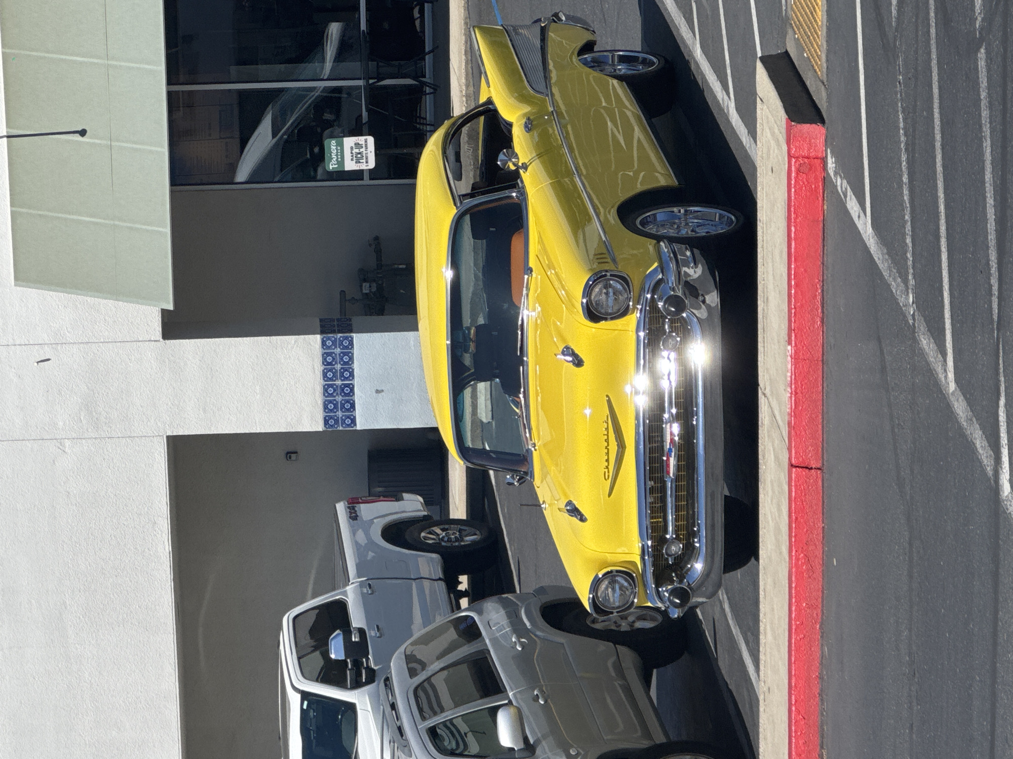 The image captures a sunny outdoor scene in Roseville, USA, featuring a vibrant yellow vintage Chevrolet parked in a lot. The classic car, a two-door coupe, is the central subject, its polished chrome gleaming under the sunlight. It is positioned prominently in the foreground, facing slightly to the right.
To the left of the vintage car, a silver SUV is partially visible, parked closely. Behind the yellow car and to the left, a white pickup truck with "4x4" badging is parked. In the background, a building with a white facade and a light green awning over a storefront is visible. A sign for "Panera Bread" with "RAPID PICK-UP" is displayed on the storefront window, indicating a commercial establishment. The ground is paved asphalt, marked with white parking lines, and a raised red curb runs along the edge of the parking area in the foreground. The overall environment suggests a commercial or retail area.