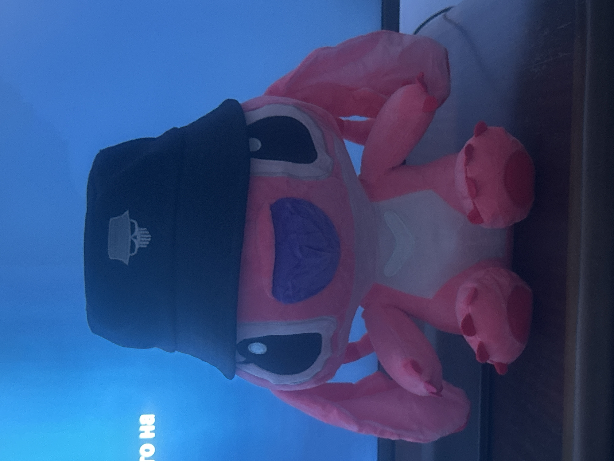 A pink plush toy, resembling the Disney character Angel from Lilo & Stitch, is centrally positioned and seated on a dark, flat surface. The plush toy wears a black bucket hat adorned with a white embroidered stylized skull emblem on the front. Its large black eyes with distinct white pupils and a purple snout are visible. In the background, a large, luminous blue screen, likely a television or monitor, displays partial white text on its left side that reads "ТО НА". No visual elements within the image provide definitive identification of its location as Pleven, Bulgaria.