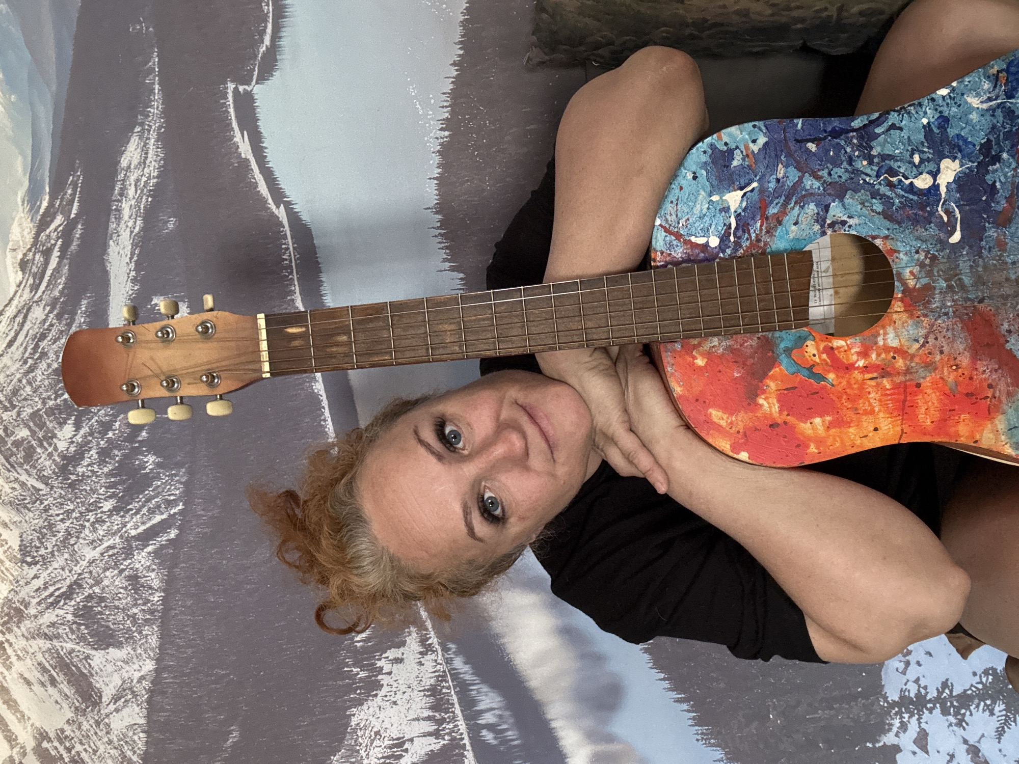 A woman with fair skin, light blonde hair, and blue eyes is lying down, cradling an acoustic guitar. The guitar has a brightly painted body, with colors including blue, orange, and white. The woman is wearing a black shirt. The background appears to be a wall with a scenic print of a mountainous landscape. There is no indication of Chaikovsky, Russia in the image.