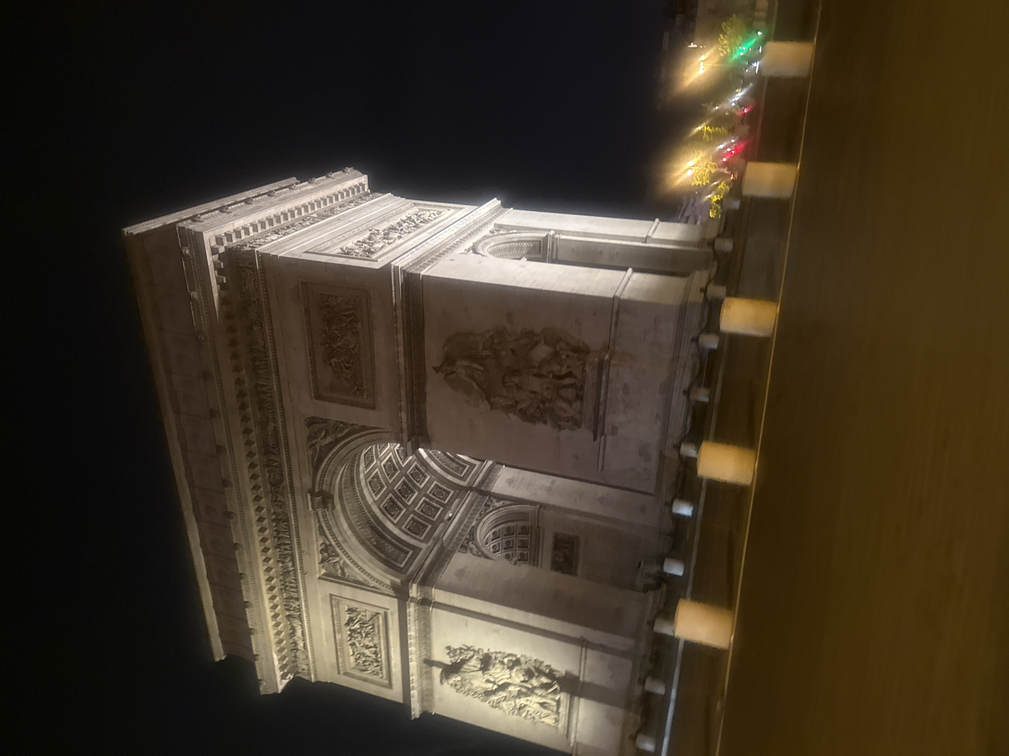 The image shows a nighttime view of the Arc de Triomphe in Paris, France. The monument is illuminated, showcasing its detailed carvings and architectural elements. Adjacent to the arch is a street, also illuminated with streetlights, with lights in the background. The sky is dark, indicating a clear night. There are no people visible in the image.