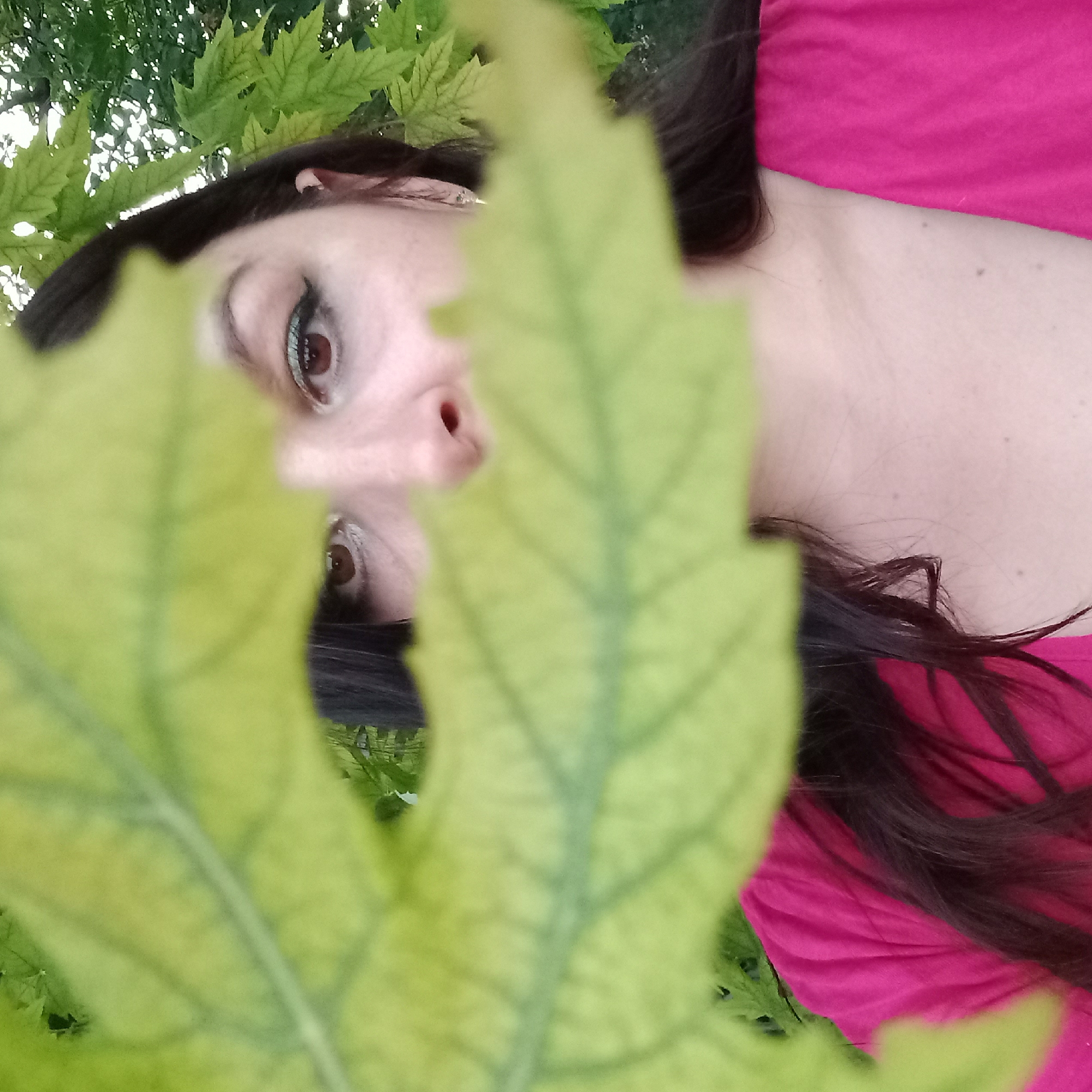 A woman with dark hair and brown eyes is partially obscured by a large, green leaf in the foreground. She is wearing a bright pink top. Her makeup includes eyeliner and what appears to be blue eyeshadow. In the background, out of focus, are green leaves and some greenery, suggesting an outdoor setting. The image was taken in Berezhynka, Ukraine.