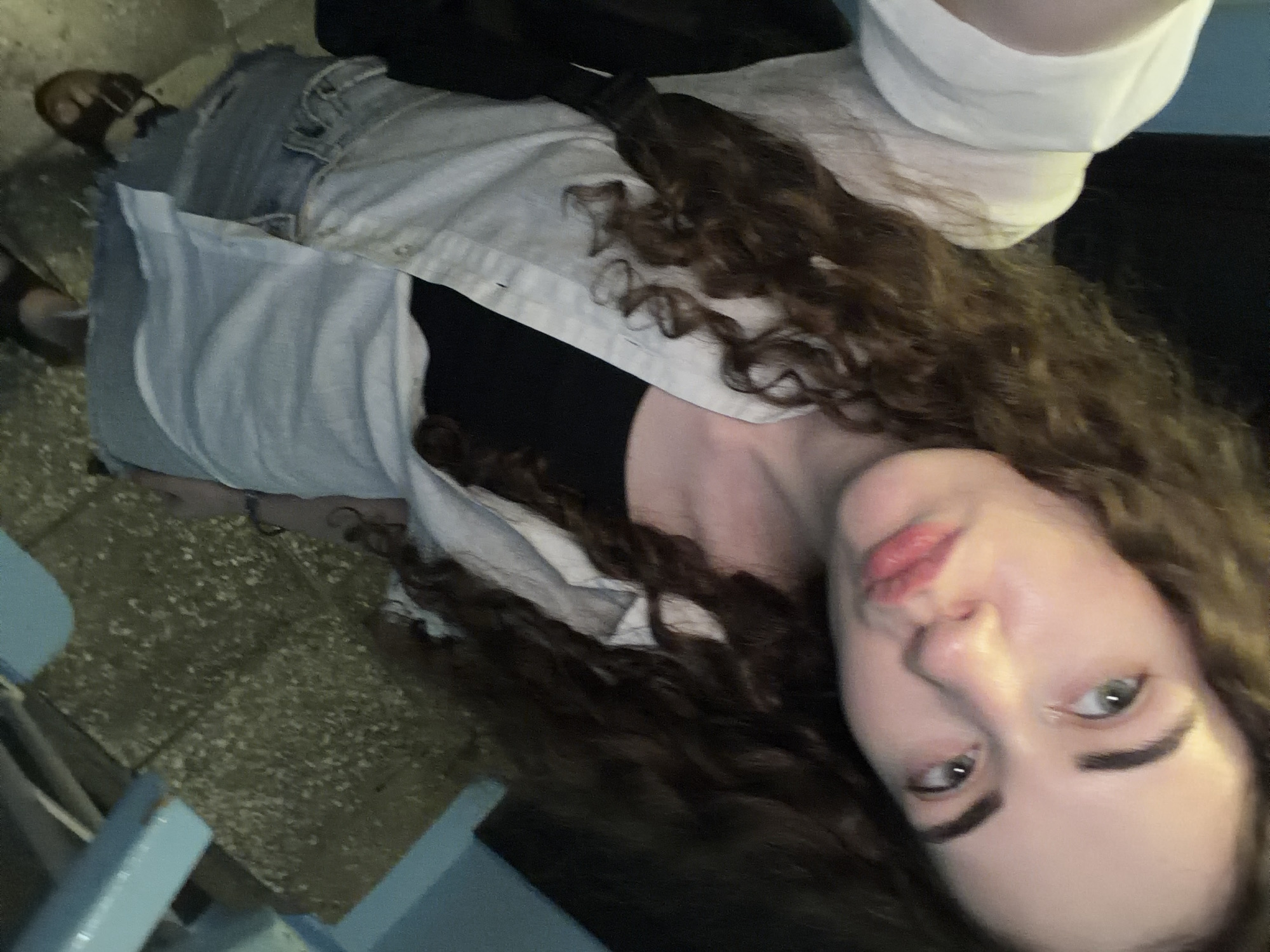 A young woman with curly brown hair and light-colored eyes is taking a selfie. She is wearing a white open button-up shirt over a black top and denim shorts. She is standing on a textured concrete surface, possibly stairs, with blue painted railings. The lighting is dim, and the background is dark. The image does not provide any specific details to determine its location in Zavodoukovsk, Russia.