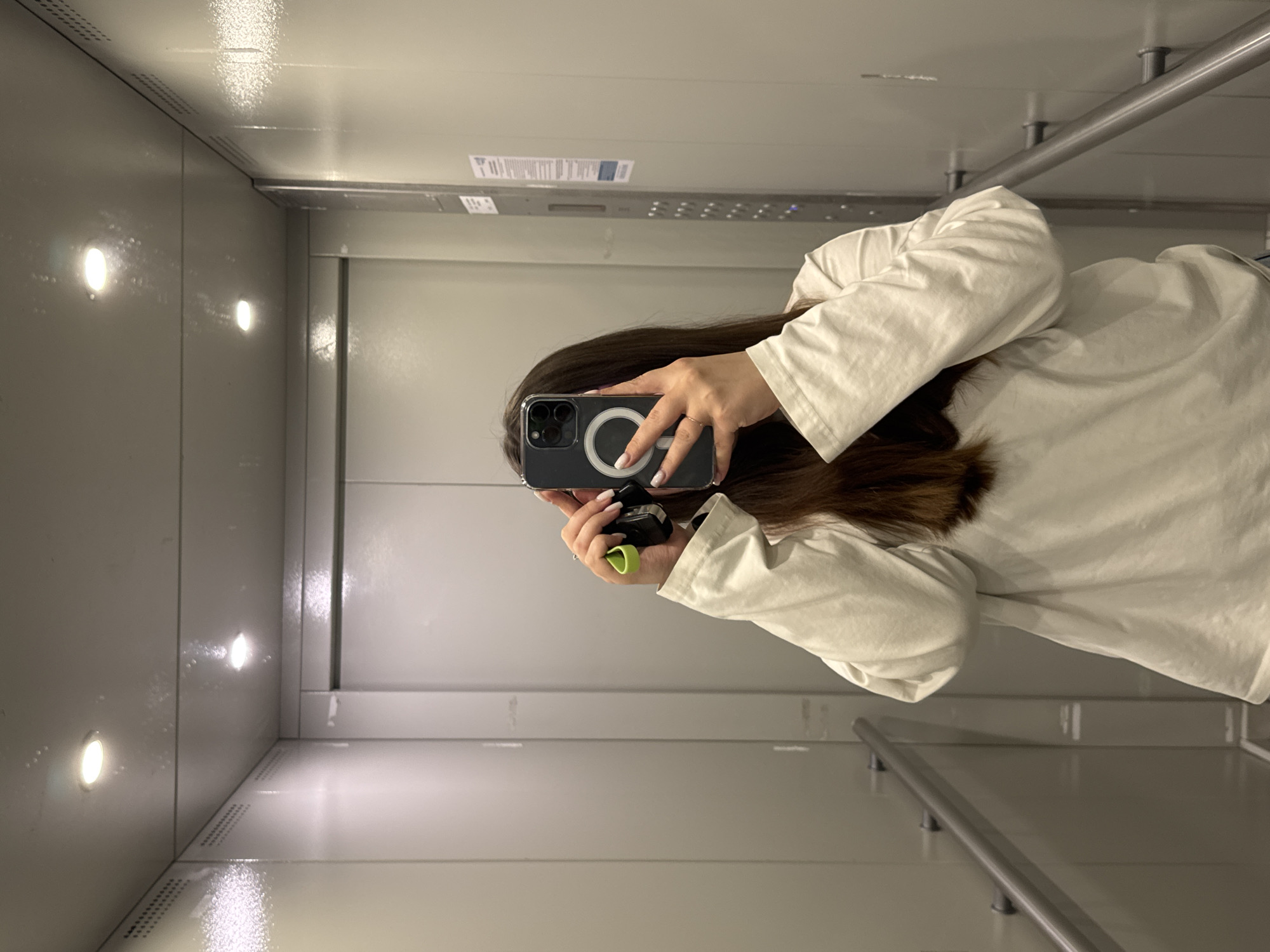 A person in a white shirt is holding a smartphone up to take a selfie inside of an elevator. The elevator's interior is made of light gray metal panels, lit by a column of round, recessed lights. A grab bar is visible on the right side of the elevator. There is a gray metal panel above the elevator doors with printed text and other markings. The location is inside an elevator. The city of Dikaya Gar', Russia is not verifiable from the image.