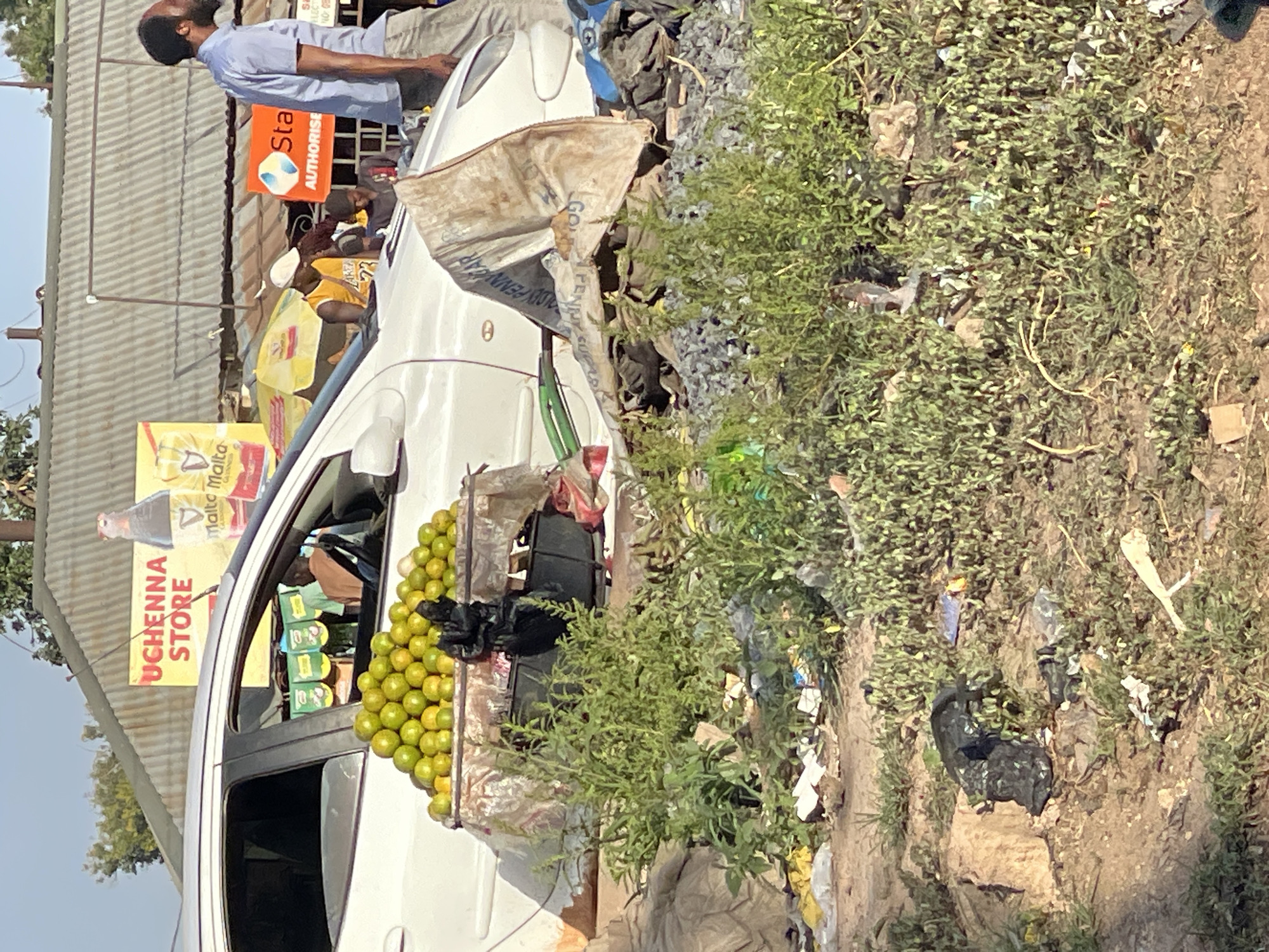 The image depicts a white car parked next to a pile of garbage in Bukuru, Nigeria. Lemons are displayed for sale from the car's passenger window. A sign reading "Uchenna Store" is visible in the background, advertising Malta Guinness. Another sign says "Sta AUTHORISE." A man in a light blue shirt stands near the car. There are other people standing around a sign in the background.