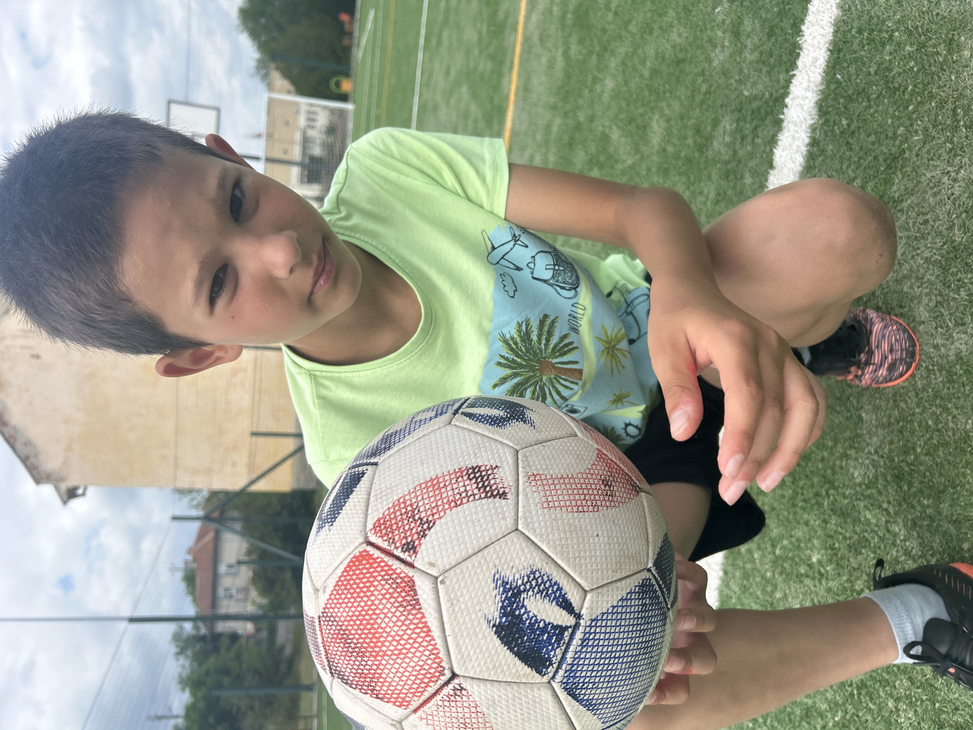 A young boy sits on a green artificial turf field, likely a soccer field, holding a soccer ball. He is wearing a light green t-shirt with a tropical-themed design and dark shorts. He has dark hair and a fair complexion. The soccer ball is predominantly white with blue and red patterned panels. The boy is looking directly at the camera with a slight smile. In the background, a building and trees are visible. The location may be in the city of Kalchevo, Bulgaria.