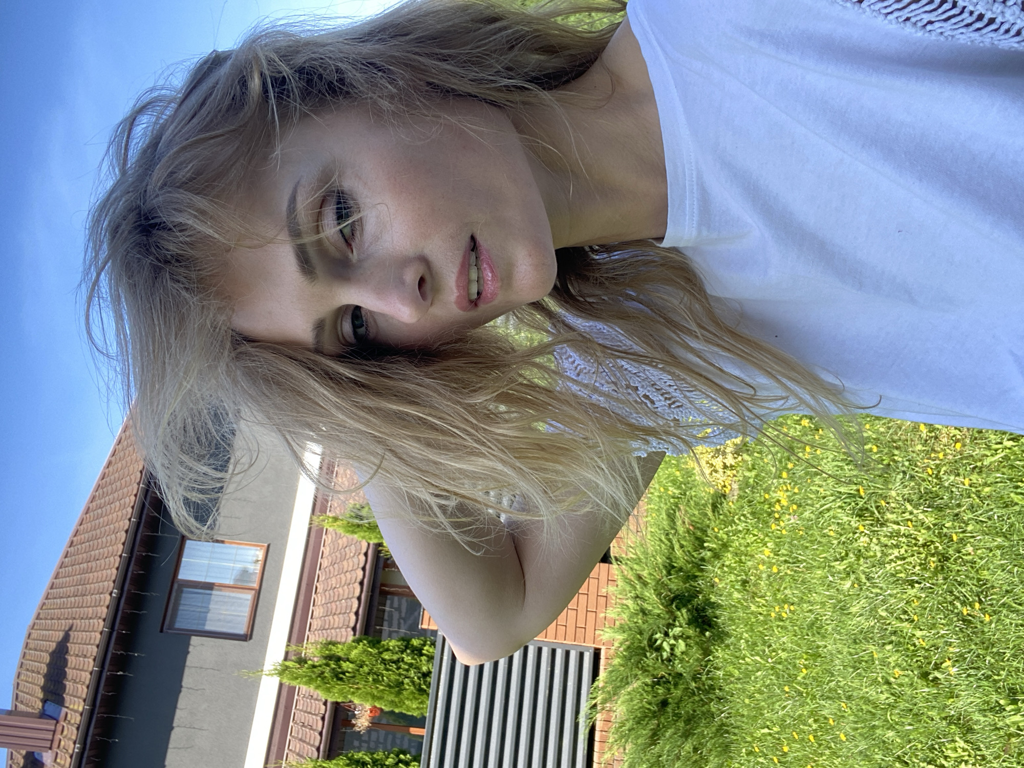 In this outdoor, eye-level, close-up selfie taken in the city of Миханавіцкі сельскі Савет, Belarus, a young adult woman with blonde, windswept hair is looking towards the camera. She is wearing a white t-shirt with textured sleeves. Her left arm is raised, with her hand resting on the back of her head. The background features a green lawn with scattered yellow wildflowers, a grey house with a brown tiled roof, and dark grey horizontal fencing. The sky above is a clear, bright blue. The woman's expression is neutral, with her lips slightly parted. The lighting suggests it is a sunny day.
