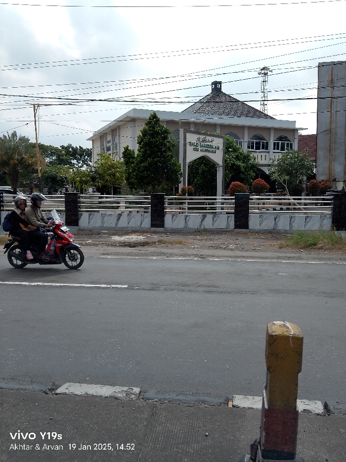 A daylight street scene in Arjawinangun, Indonesia, captures a red motorcycle with two occupants driving along an asphalt road. The driver wears a helmet and a light-colored jacket, while the passenger, also helmeted, carries a backpack and is seated behind.
In the background, a prominent building, likely a mosque, is visible behind a fence with marble-patterned panels and dark pillars, and lush greenery including trees and shrubs. The building features a distinctive multi-tiered, pyramidal roof structure. A large sign in front of the building displays text in Arabic script, which reads "Masjid Baabus Salaam," and in Latin script "KREO ARJAWINANGUN," identifying it as a mosque. To the right of the mosque, a lattice communication tower stands tall. Utility lines crisscross the upper part of the frame against an overcast sky.
In the foreground on the right, a yellow and orange painted concrete post is partially visible. The road itself appears dark and slightly wet or freshly paved. The image includes a watermark at the bottom left indicating it was taken with a "vivo Y19s" by "Akhta & Arvan" on "19 Jan 2025, 14:52" (2:52 PM).