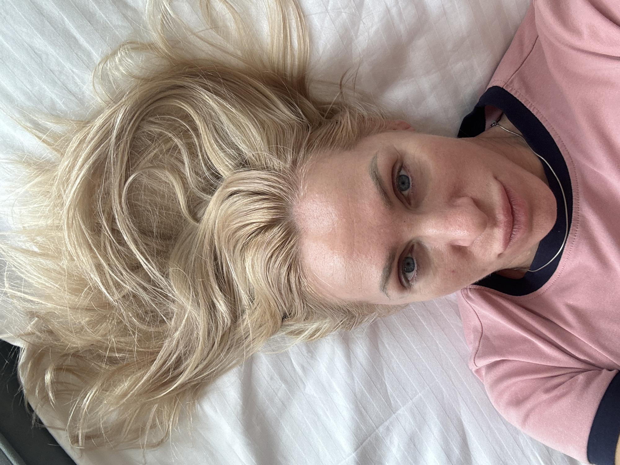 A blonde woman lies on her back on a white, ribbed surface, possibly a bed. Her head is centered in the frame, with her long, blonde hair fanned out above her. She is wearing a pink t-shirt with a dark blue collar and trim. Her eyes are open and looking at the camera, and her lips are slightly parted. The lighting suggests an indoor setting, possibly a bedroom. There are no visible indications of the city of Starye Chernitsy, Russia.
