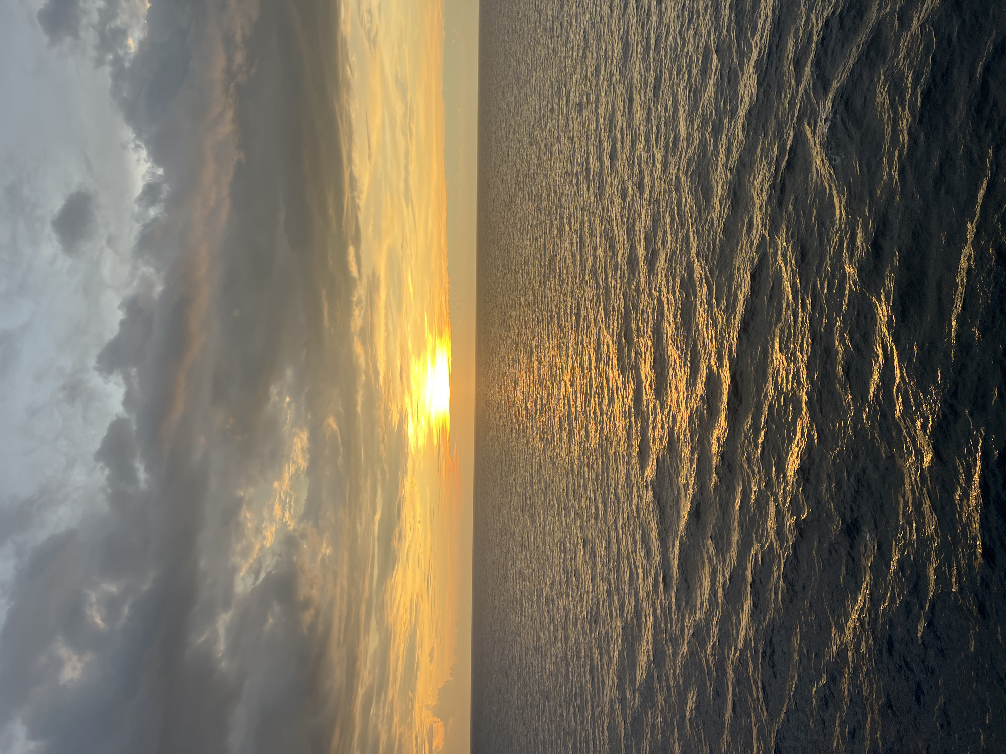 A wide-angle shot captures a serene seascape during either sunrise or sunset. The sun, a bright orb of yellow-white light, is positioned just above the horizon, casting a golden pathway across the rippling water. The sky above is a gradient of warm hues, transitioning from soft yellows and oranges near the sun to muted grays and blues further up, with scattered clouds adding texture. The ocean is vast and calm, with gentle waves reflecting the warm light of the sun. The perspective is from a high vantage point, likely a ship or elevated structure, looking out towards the open water. There are no people or man-made structures visible in the frame, emphasizing the natural beauty of the scene.