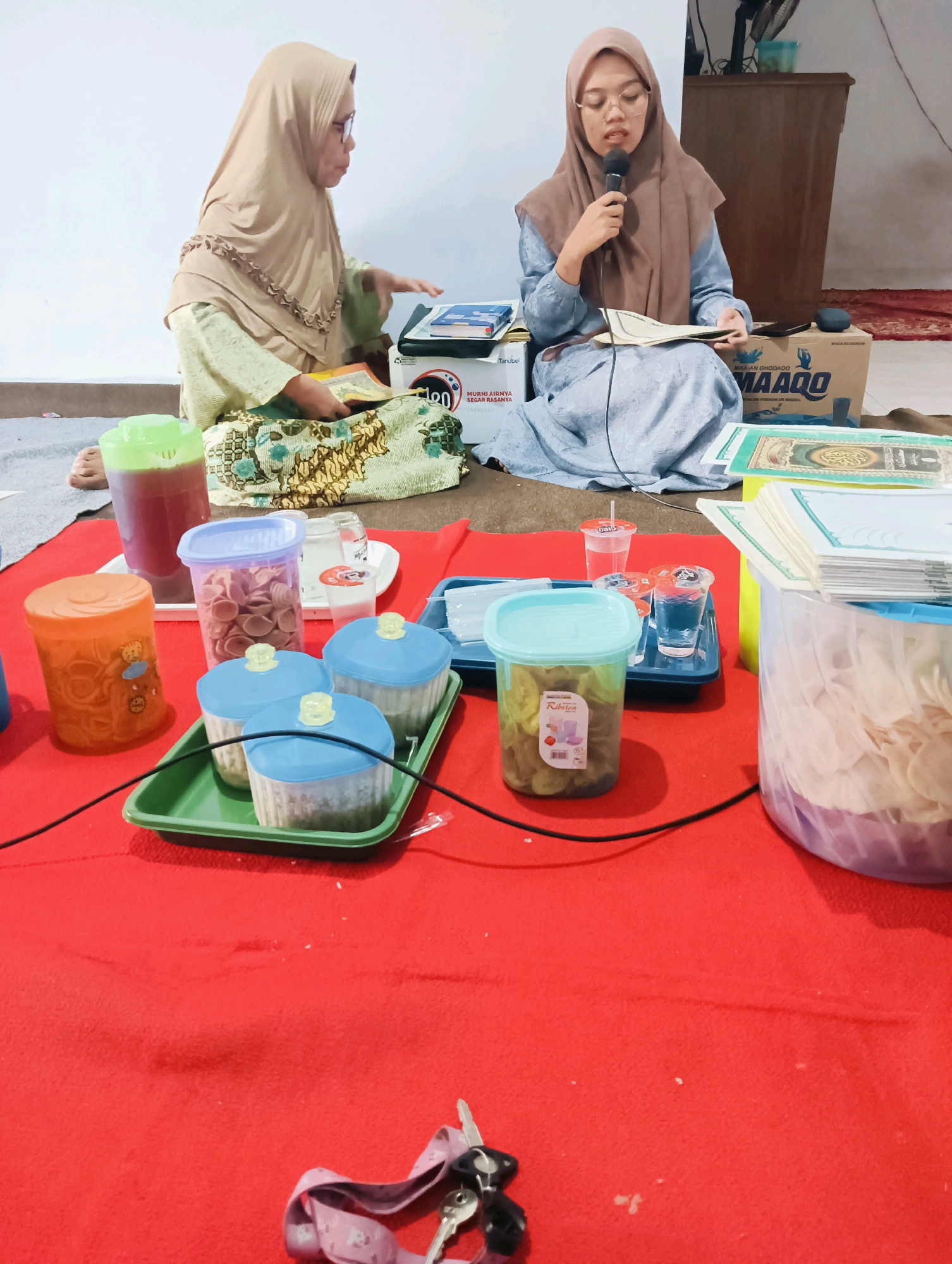 In the city of Jatigedong, Indonesia, two individuals are seated on the floor, engaged in what appears to be a religious or educational activity. The person on the right, wearing a brown hijab and glasses, is holding a microphone and speaking, while also holding a book. The person on the left, also wearing a hijab, is looking towards a book or papers placed on their lap, and appears to be gesturing with their hand.
The environment is a room with a white wall in the background. A wooden cabinet or furniture is visible behind the individuals. The floor is covered with a red fabric, likely a carpet or blanket, on which the people are sitting. In the foreground, several plastic containers and cups are arranged on the red surface. These containers hold various items, including what looks like snacks, drinks, and possibly religious texts. One container has a green lid and holds a reddish liquid, another has a purple lid and contains light-colored snacks. There are also several clear plastic cups with drinks, some with straws.
A prominent cardboard box with "MAAQO" branding is visible in the background, further suggesting a commercial or organized event. The overall scene suggests a gathering or lesson, possibly related to Islamic studies or recitation, given the presence of books and the attire of the individuals. The lighting appears to be artificial, contributing to the indoor setting. A bundle of keys with a pink lanyard lies on the red surface in the bottom right of the frame.