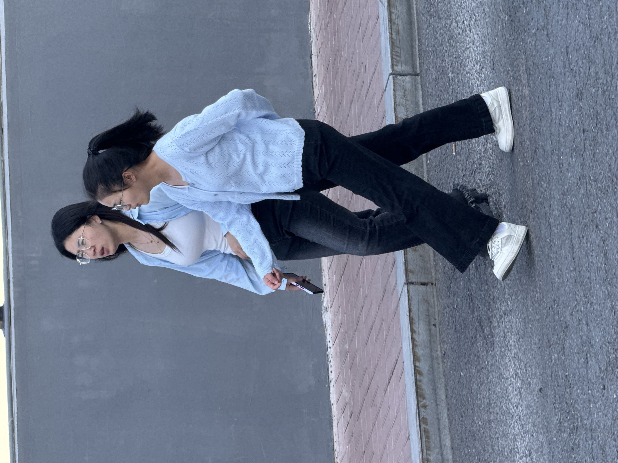 Two young women stand together on a paved surface with one arm around the other. They wear jeans and white tennis shoes. One has on a white shirt and light blue sweater. The other has on a darker shirt. They both wear eyeglasses. One woman holds a smart phone. The backdrop includes a gray wall and concrete and brick. The scene is consistent with an urban area. The city of Zhongtun, China, cannot be determined from the visual elements provided.