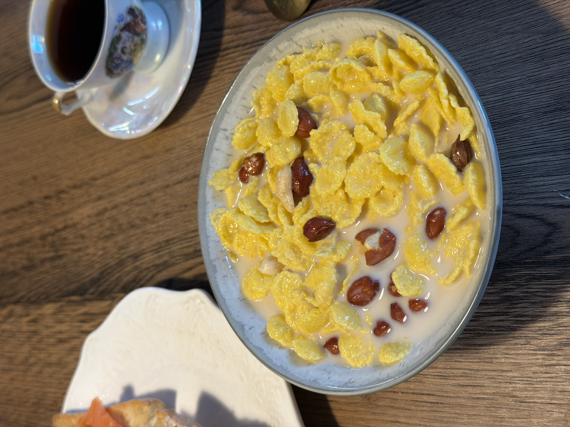 A close-up, eye-level shot shows a bowl of cornflakes with milk and peanuts on a wooden table. To the left, a white plate holds a sandwich with smoked salmon. In the background, a teacup filled with dark liquid sits on a saucer. The setting is a room in the city of Perm, Russia, with warm lighting and a rustic ambiance.