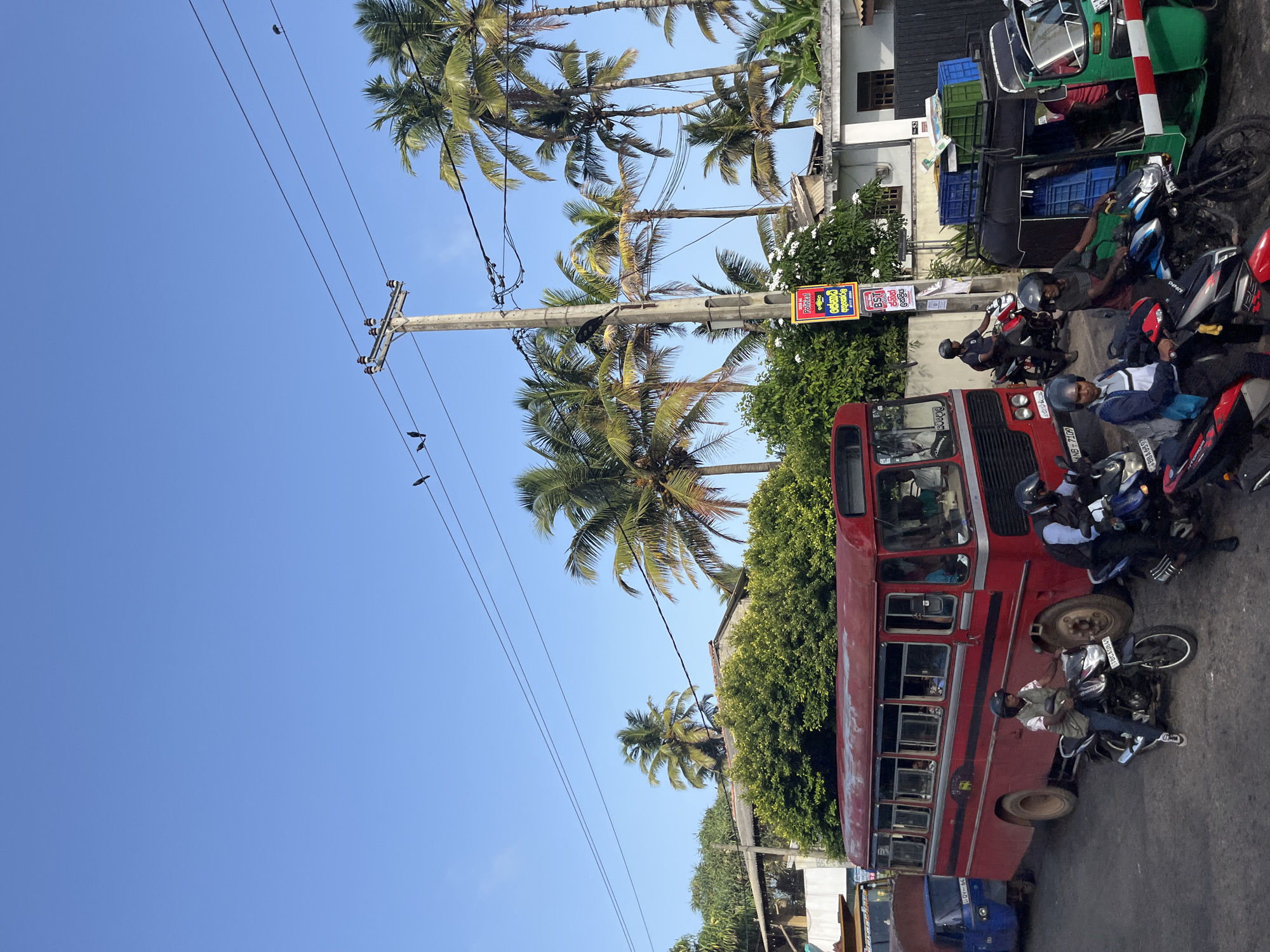 The image captures a street scene in Maggona, Sri Lanka, under a clear blue sky. A red bus is parked on the right side of the road, with several people visible inside. In front of the bus, a man on a motorcycle waits. To the left of the bus, a blue three-wheeled vehicle is also present. Several other motorcycles and riders are scattered throughout the scene, indicating traffic. Palm trees line the background, suggesting a tropical or subtropical environment. A utility pole with electrical wires stands in the middle of the frame, with a few birds perched on the wires. A red and white barrier is visible in the lower right corner, and several blue and green crates are stacked on top of a vehicle. A political poster is attached to the utility pole, featuring text in a foreign language. The overall impression is that of a busy street with mixed traffic and local elements.