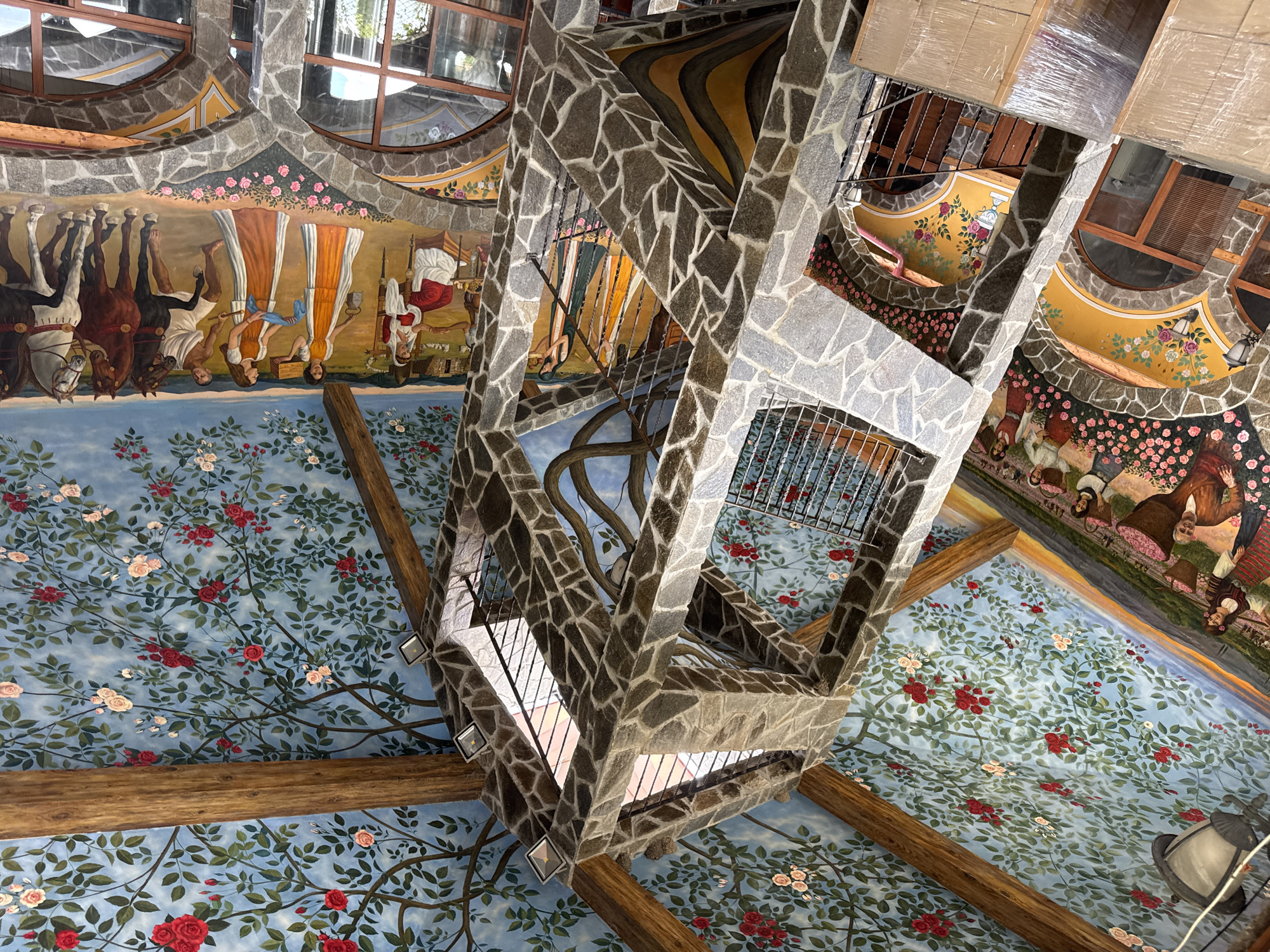 The image depicts an upward view within a building located in Skobelevo, Bulgaria. The ceiling is ornately decorated with a blue background and a pattern of rose bushes. Along the upper walls are detailed frescoes depicting historical figures and scenes. A central, multi-tiered structure made of rough-hewn stone ascends through the center of the frame. This structure features metal railings at each level. Wooden beams intersect the painted ceiling, adding structural support. Windows are visible near the top of the image, allowing natural light to enter. Overall, the scene portrays a building with unique architectural and artistic elements.