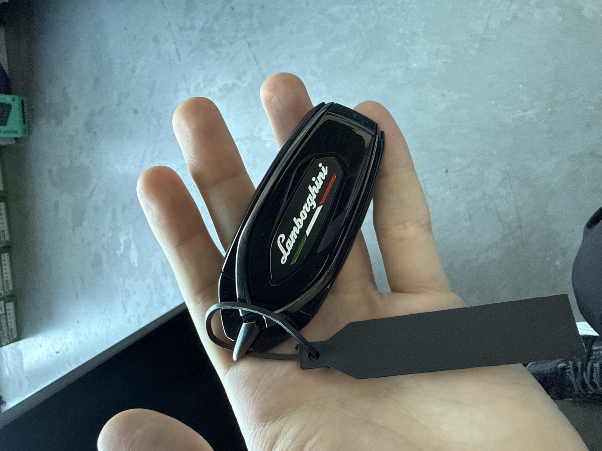 A person's hand holds a black Lamborghini car key with a black tag attached. The background appears to be a light gray surface, possibly a desk or countertop, with some dark objects visible in the periphery. The Italian flag colors are incorporated into the Lamborghini logo on the key. No information about the city of Plovdiv, Bulgaria is discernible.