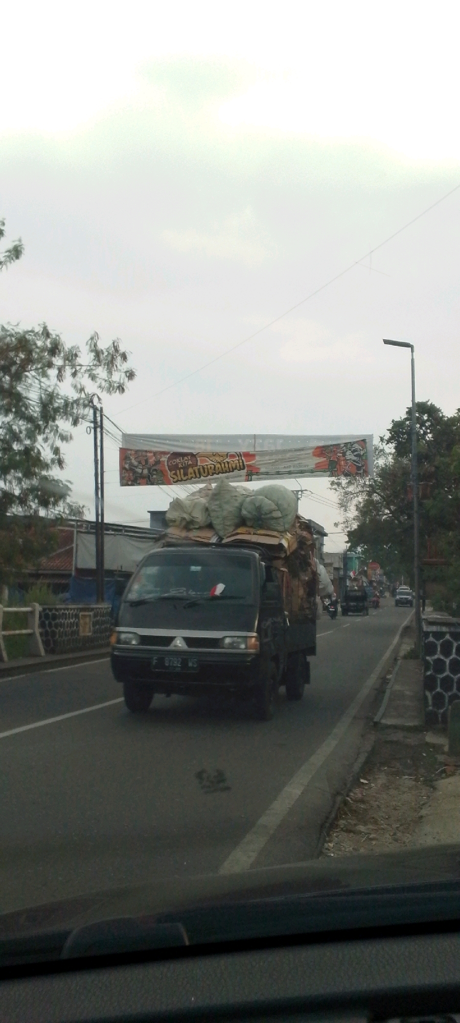 A black Mitsubishi L300 pickup truck with its bed heavily loaded with cargo, including large sacks, is driving on a paved road. The truck's license plate reads "F 8792 WS." A red and white Indonesian flag is visible on the truck's dashboard. The truck is in motion, as indicated by the blur of the surrounding environment and the road ahead. The scene appears to be in an urban or semi-urban area, with buildings and trees visible on the sides of the road. A large banner hangs over the road with text that includes "SILATURAHMI." The sky is overcast, suggesting a cloudy day. The image was taken from inside another vehicle, as indicated by the view of a dashboard in the foreground. The location is identified as Cisalak, Indonesia.