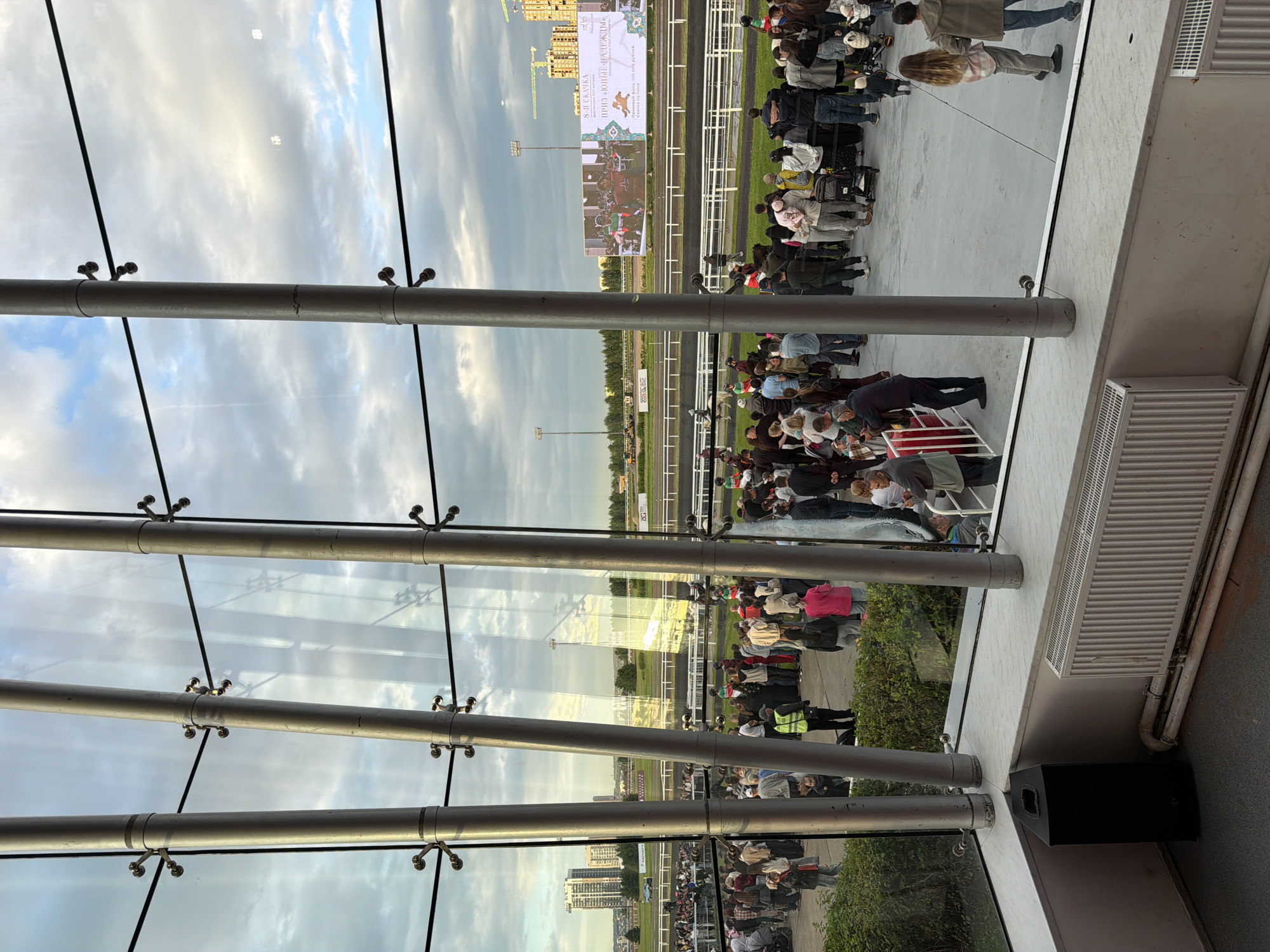 The image captures an indoor view looking out onto a horse racing track. Several large, vertical metal poles and horizontal black wires are prominent in the foreground, acting as structural elements of the building and framing the scene outside. Through a large glass window, a crowd of people is gathered on a paved area next to the track, some appearing to be spectators. A horse race is visible on the track, with jockeys in colorful attire. A large digital screen displays images related to the event, including horses and text in Russian. In the background, buildings and construction cranes indicate an urban environment. The sky is partly cloudy with a mix of blue sky and grey clouds. Several radiators and a black speaker are visible indoors, near the glass partition.