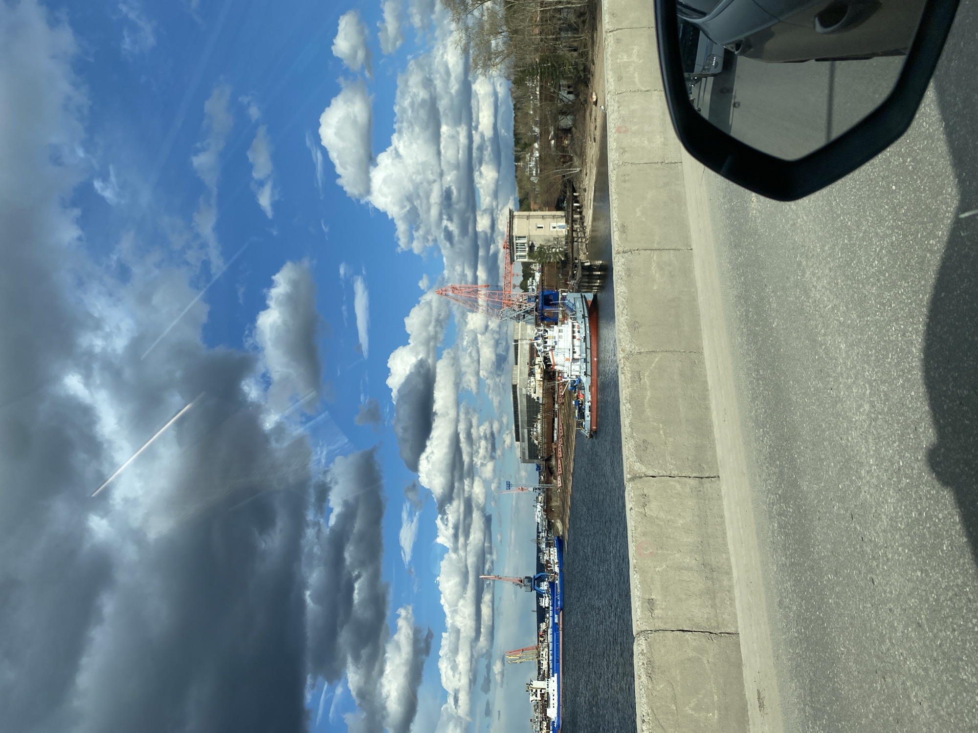 The image captures a vibrant, partly cloudy day over an industrial waterfront from the perspective of a vehicle on an elevated road or bridge. The scene is dominated by a wide expanse of blue sky dotted with puffy white clouds, contrasting with some darker, grey clouds. Several bright white contrails from aircraft are visible in the sky.
In the foreground, a concrete barrier and a section of asphalt road are visible, leading to the car's side mirror, which occupies the upper right portion of the frame (when upright). The mirror reflects the dark exterior of the car and the road behind it.
Below the road, a dark body of water, likely a river, stretches towards the horizon. Along the opposite bank, several large ships and barges, some white and blue, are docked. The waterfront is an active industrial area, featuring multiple tall, orange-red gantry or port cranes. Behind these industrial structures, various buildings, including what appears to be a multi-story building, are visible, interspersed with trees, many of which are bare, suggesting a late winter or early spring setting. Further into the background, a residential or forested area can be seen.
The activity depicted centers on the port operations, with ships moored and cranes positioned for loading or unloading. The time of day appears to be daytime with clear visibility, and the general atmosphere is one of industrial productivity against a dynamic sky. No discernible text is visible on any objects, buildings, or vehicles. The location context indicates this scene is in Chaikovsky, Russia.