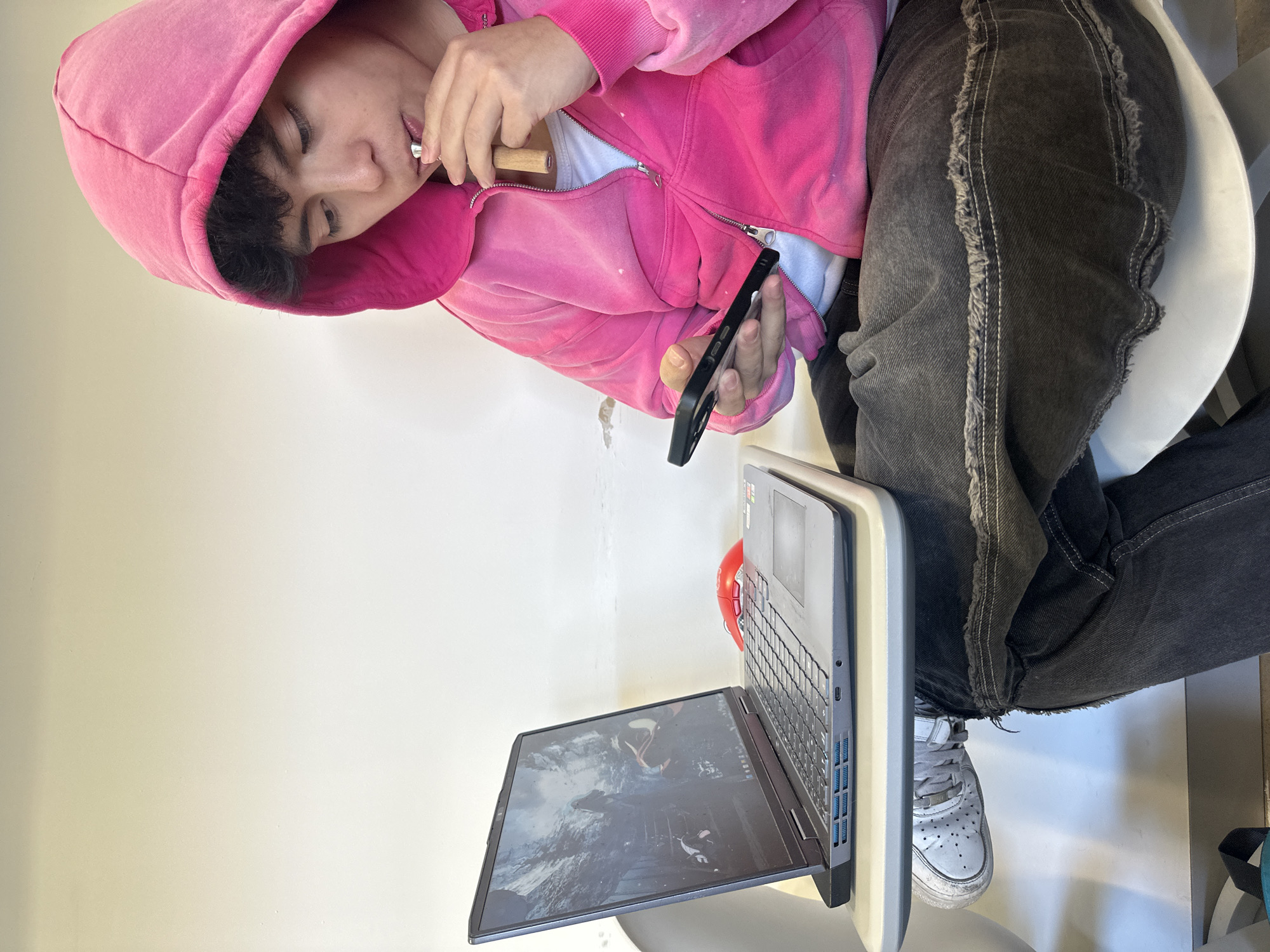 A person wearing a pink hoodie and grey jeans sits in a white chair with a laptop open in front of them. They are holding a black smartphone in one hand and a wooden and metal object near their mouth with the other. The laptop screen displays a dark image, potentially from a video game. The background is a plain white wall with some minor imperfections. The location cannot be determined as Mexico City, Mexico.