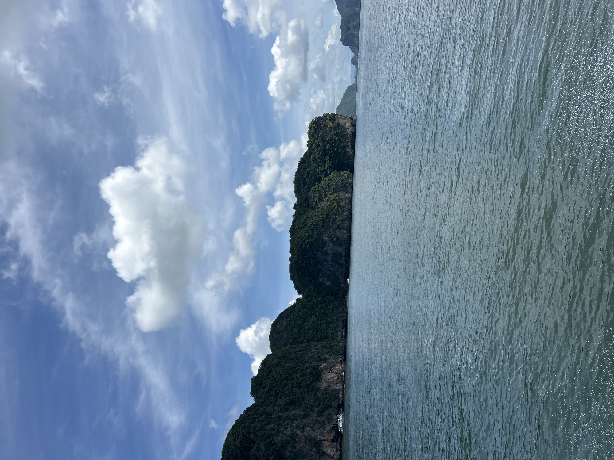 A serene view of Phang Nga Bay, Thailand, showcases dramatic karst islands covered in lush green vegetation rising from the calm, blue-green waters. The sky above is a vibrant blue, adorned with scattered white cumulus clouds, some appearing wispy and others more voluminous. The water's surface is gently rippled, reflecting the light and creating a shimmering effect. In the distance, several boats are visible, including a larger white vessel with a blue stripe near the shore of one of the islands, and smaller boats further out. The overall scene evokes a sense of tranquility and natural beauty.