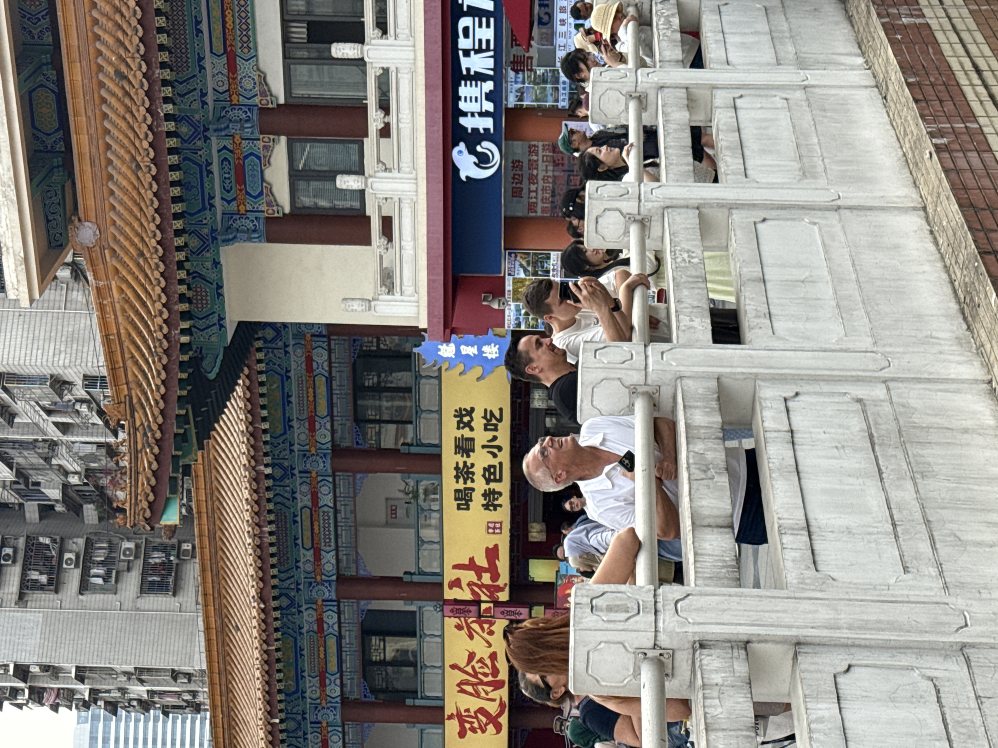 This image shows a group of people looking over a traditional marble railing in Yuzhong District, China. To their right, there is a building adorned with Chinese characters, signage, and ornate architectural details including a traditional Chinese-style roof with wooden tiles. In the background, modern buildings are visible. The scene has a blend of traditional Chinese architecture juxtaposed with contemporary urban elements. Signage for "C Trip" is visible.