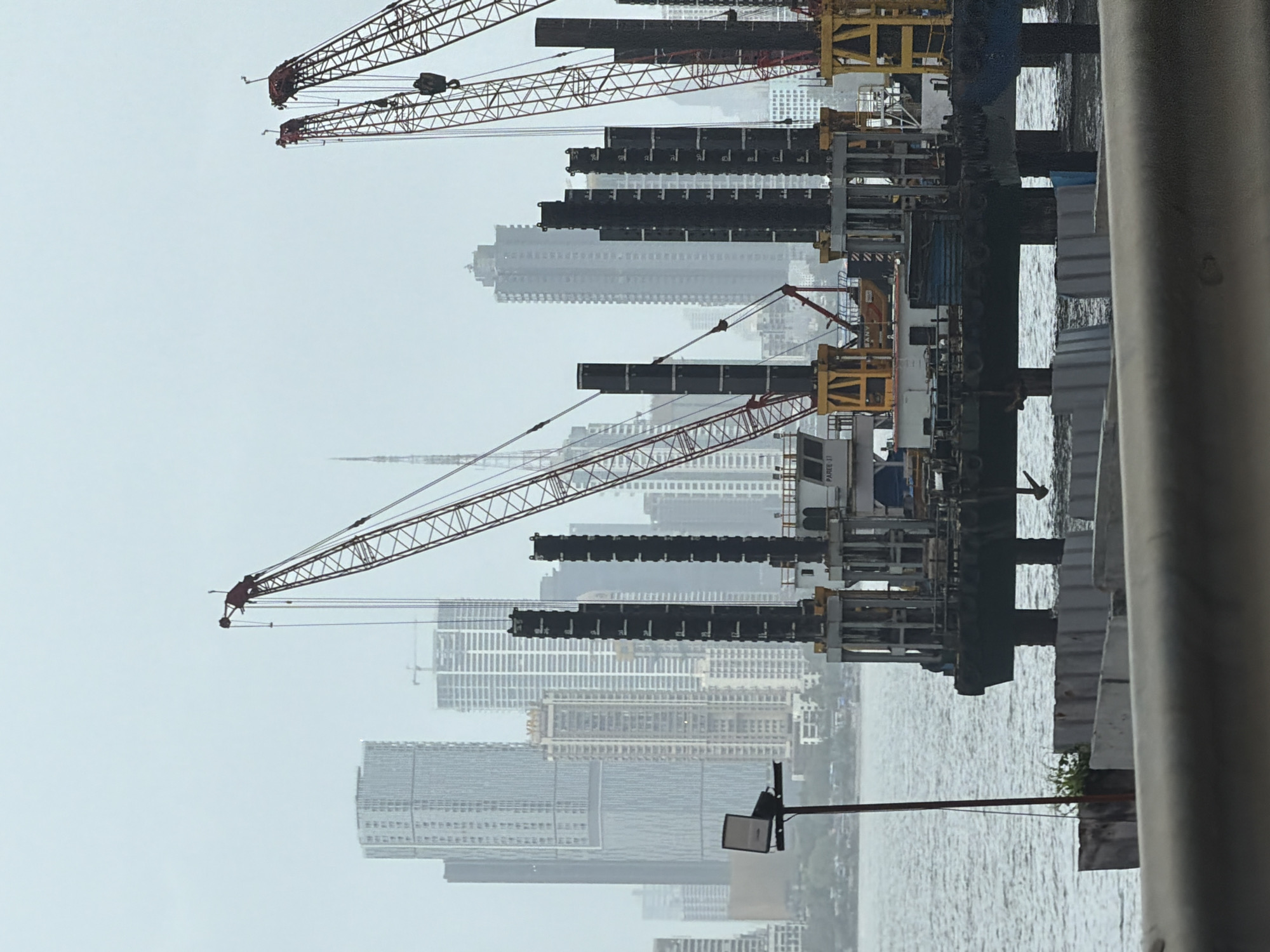This is a photograph of a construction site in Mumbai, India, likely along the coast or a river. In the foreground, there are concrete barriers and the edge of a road or walkway. The middle ground is dominated by a large construction platform with multiple cranes, pylons, and what appear to be pilings being driven into the water. The platform is supported by large pillars extending into the water. In the background, a dense skyline of tall buildings, typical of Mumbai, rises under a hazy, overcast sky. The overall scene suggests ongoing marine construction work in an urban environment.