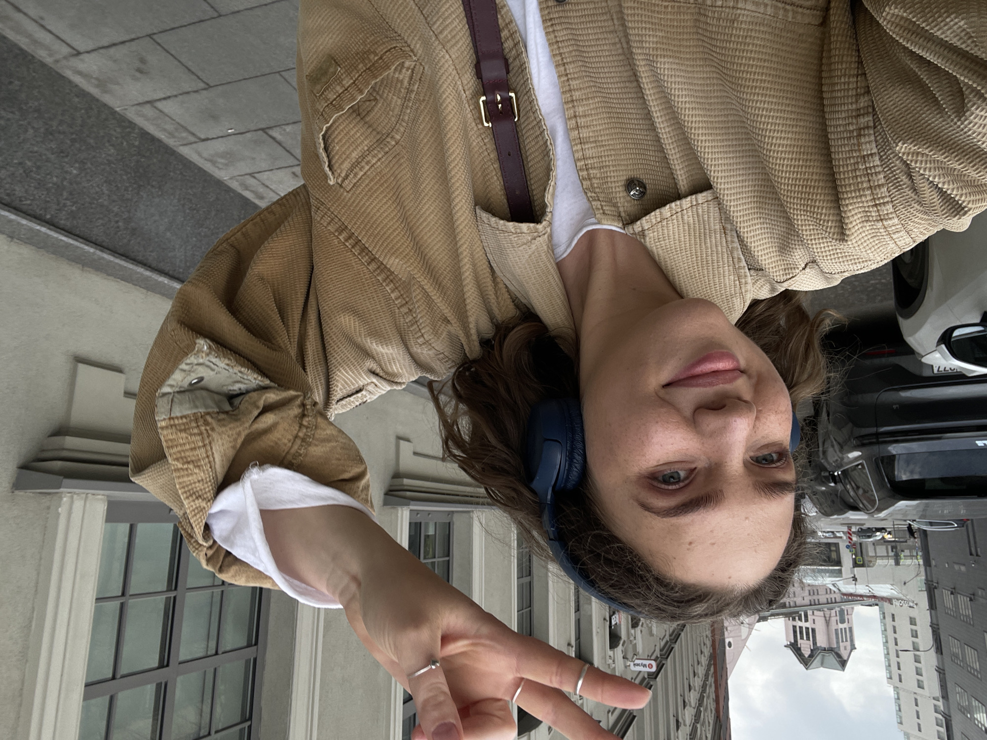 A young woman takes a selfie on a Moscow street. She wears blue headphones, a tan corduroy jacket, and a white t-shirt, and she makes a peace sign with her hand. Buildings and a car are visible in the background, as well as street signs in Russian. The street is a busy urban area.
