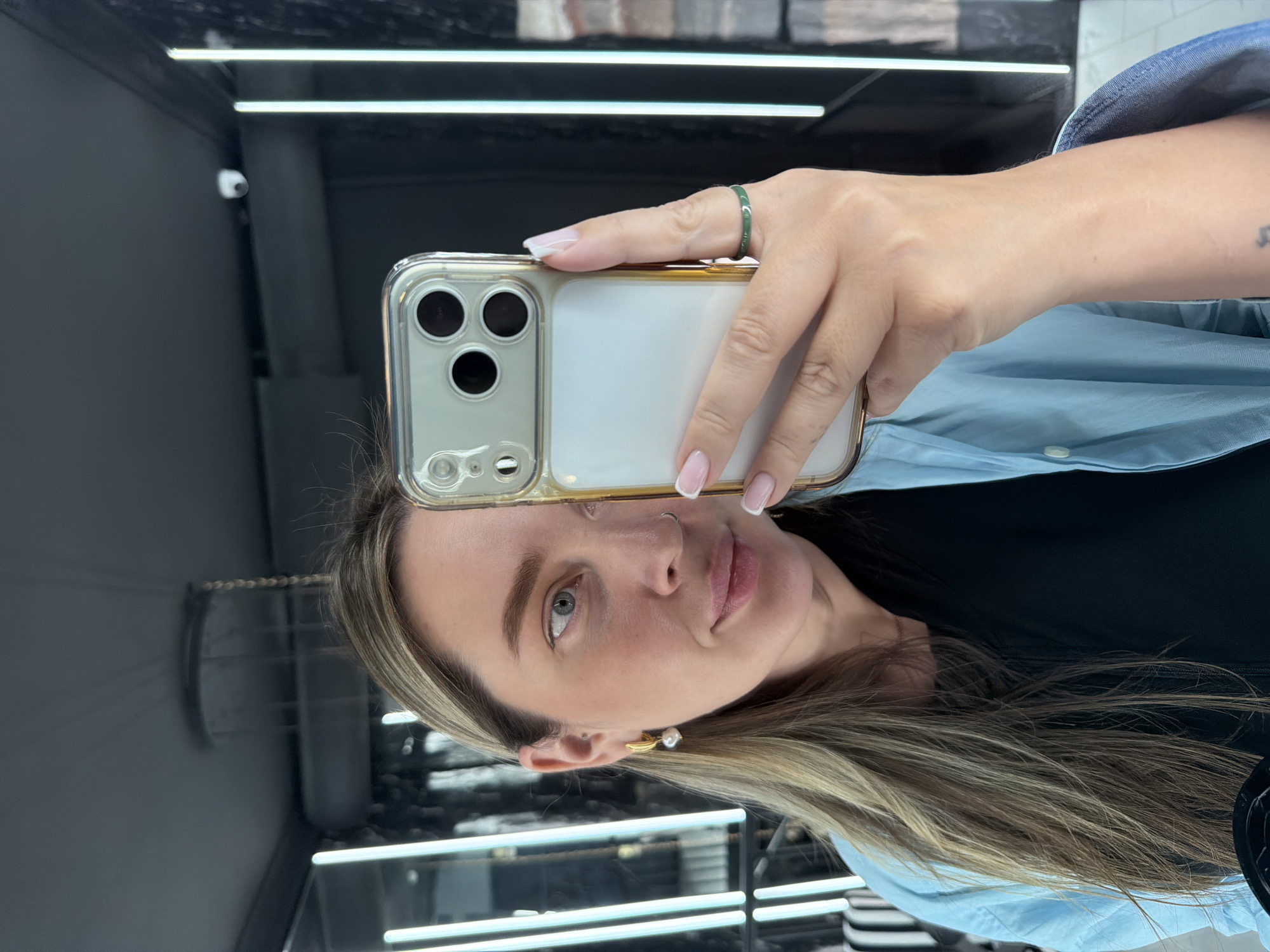 A woman is taking a selfie with her smartphone, likely in an indoor setting. She is holding the phone in her right hand, and the camera is facing her. She has blonde hair, fair skin, and is wearing a blue shirt over a black top. She is wearing a green ring on the index finger of her right hand and a gold and pearl earring in her left ear. The background appears to be a store or salon with reflective surfaces, bright lights, and some dark-colored fixtures. There is a visible brick-like pattern on a wall.
There is insufficient information to determine the specific location as Moscow, Russia, from this image alone.