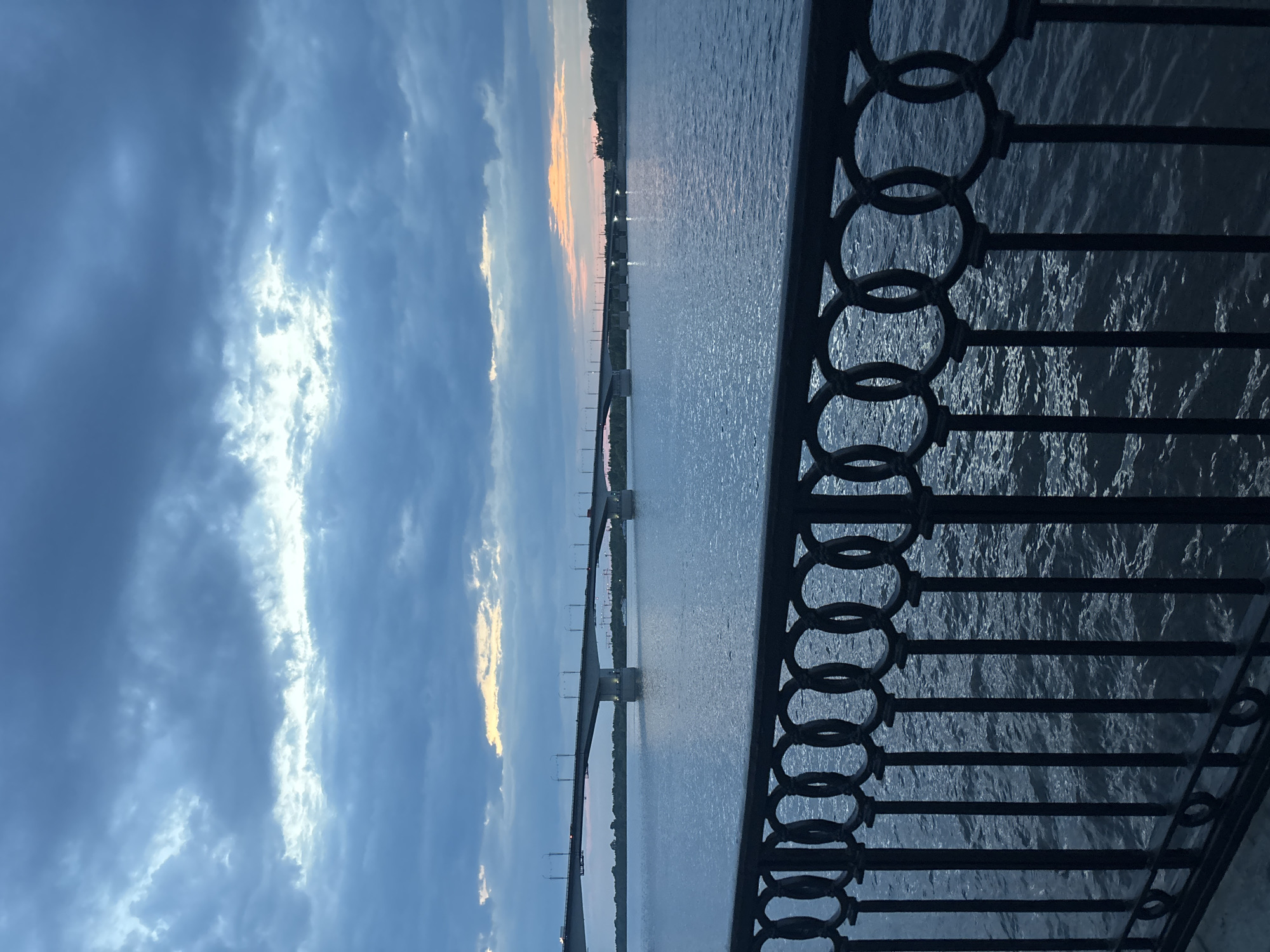 The image depicts a cityscape in Perm, Russia, at what appears to be dusk or dawn. A metal fence with circular and linear elements dominates the foreground on the right side of the frame. Beyond the fence, a large body of water reflects the sky. A bridge with multiple supports spans the water. The sky is filled with clouds, showing light breaking through, indicating either a sunrise or sunset. There are no people visible in the frame.