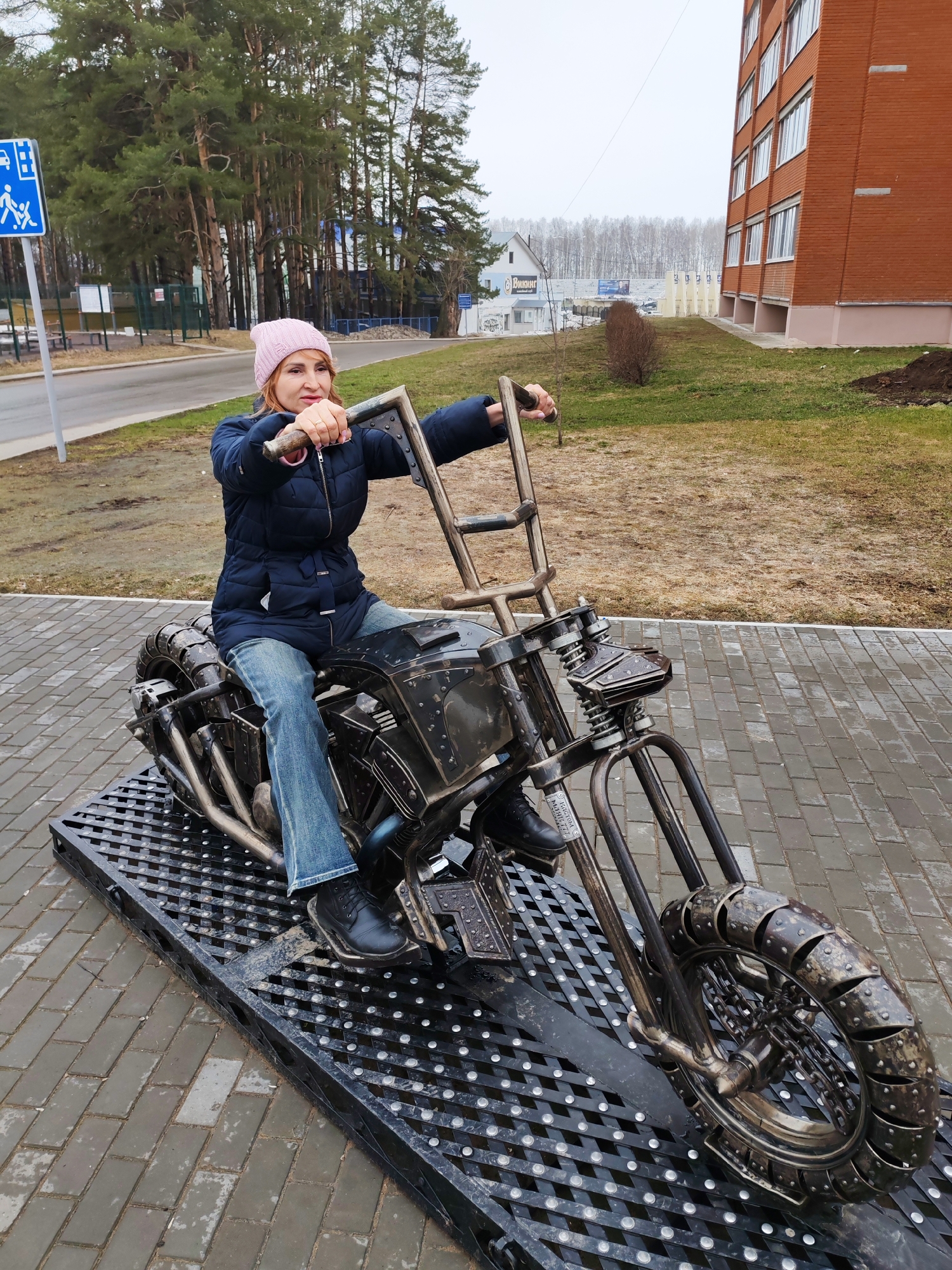 A woman wearing a pink hat, a dark blue quilted jacket, and jeans is sitting on a metal motorcycle sculpture. The motorcycle is mounted on a black metal platform with a grid pattern, which is placed on a paved area. The background includes trees, a road with a pedestrian crossing sign, a brick building, and grassy areas. This scene is located in the city of Chaikovsky, Russia.