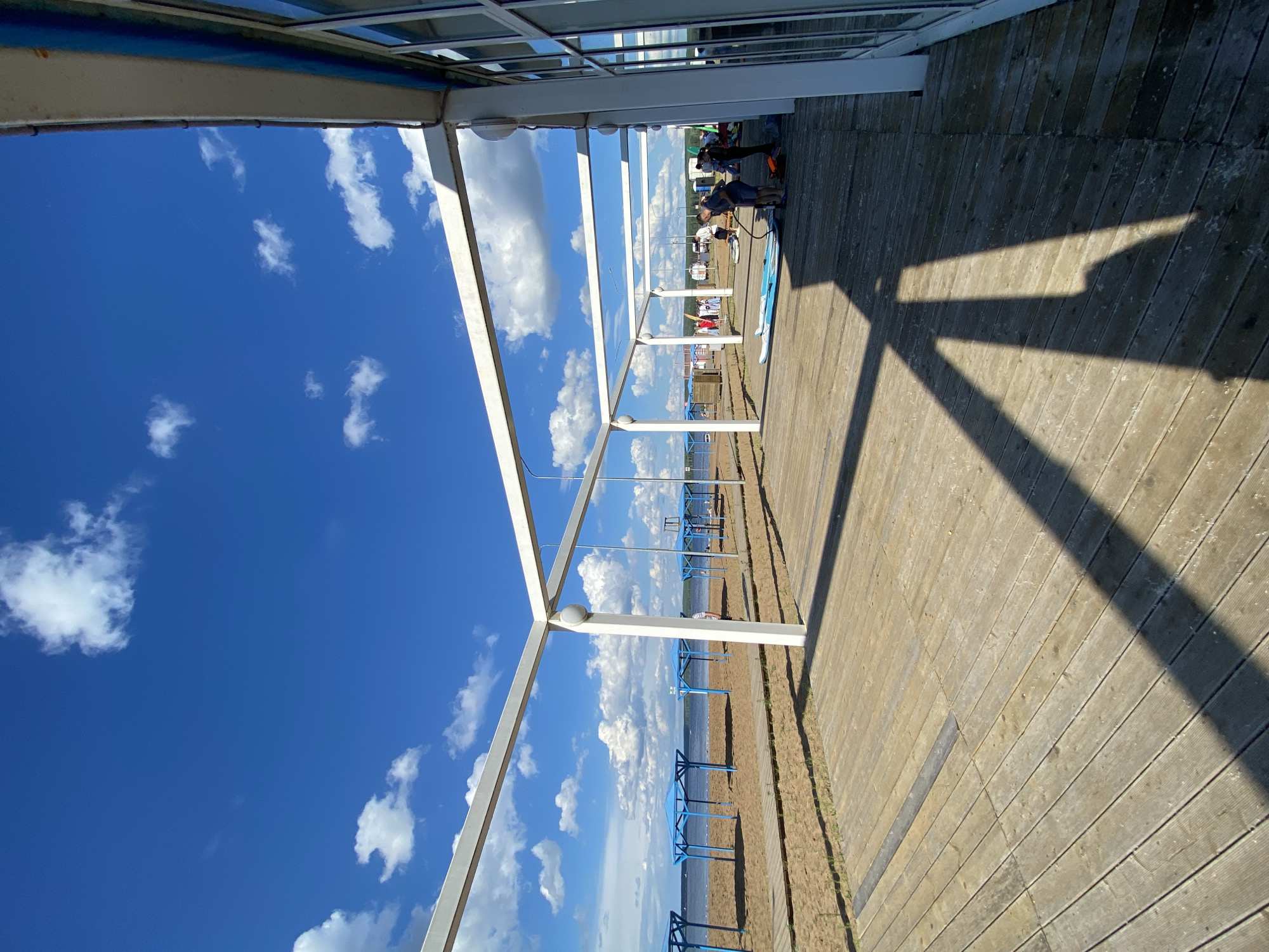 An outdoor scene on a sunny day at a beach in Chaikovsky, Russia, featuring a wooden deck in the foreground under a bright blue sky with scattered white clouds. A white canopy structure extends over the deck. To the left, a sandy beach is visible with several blue-covered shelters and pathways leading to the water. In the middle ground, a group of people are gathered near some paddleboards, some of whom appear to be preparing for or engaging in watersports. The overall environment suggests a recreational area by a body of water, possibly a lake or river, given the presence of the beach and calm water.