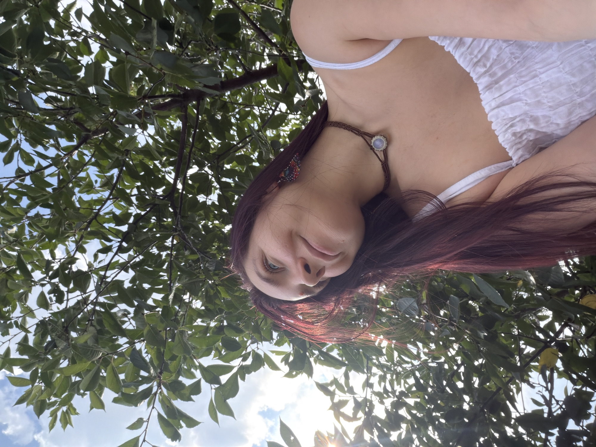 The image is an outdoor, low-angle, waist-up shot of a young woman looking up and to the left. She has long, dark red hair and is wearing a white, ruffled top and a brown macrame necklace with a pale blue stone. She is also wearing a colorful, butterfly-shaped earring in her right ear. The background consists of green leaves and branches of a tree, with glimpses of a blue sky and white clouds. Sunlight is filtering through the leaves, creating a lens flare on the left side of the image. The scene appears to be in a park or natural setting within the city of Fokino, Russia.