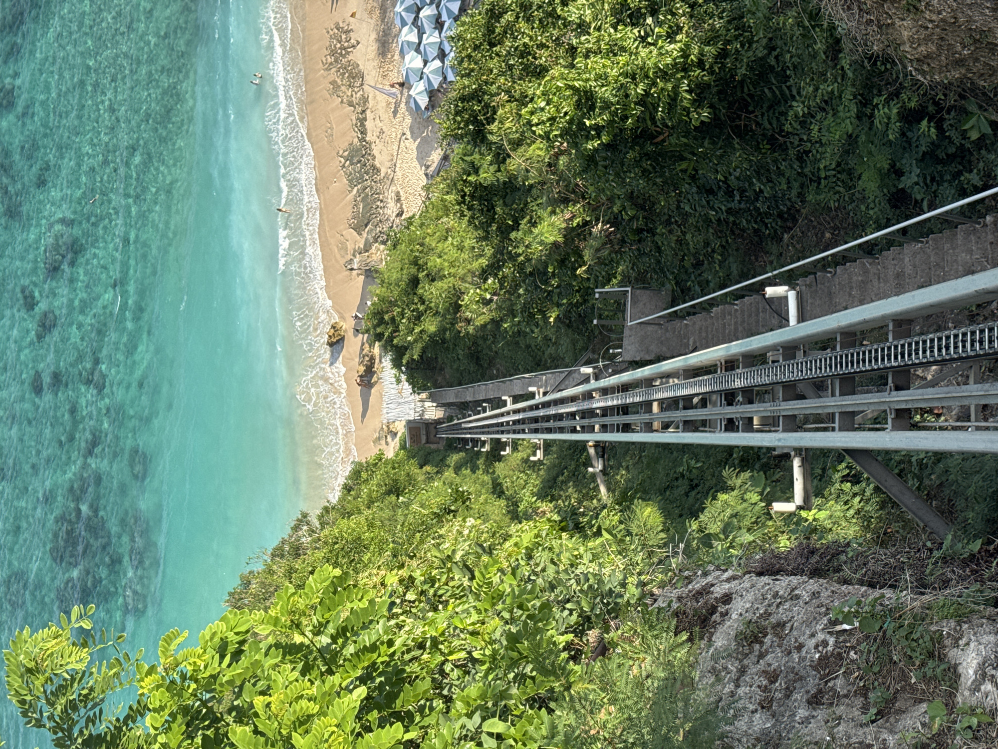The image captures a dramatic view from a cliff overlooking a beach and the ocean in Kuta Selatan, Indonesia. A metal funicular track descends steeply down the cliff face, flanked by lush green foliage. To the right of the funicular, a concrete staircase runs parallel. Below, the sandy beach is dotted with rocks and features a row of white and green striped umbrellas. The turquoise ocean extends to the horizon, with visible coral reefs beneath the clear water. Several people are present on the beach and in the water, enjoying the tropical setting. A small yellow vehicle is parked near the edge of the beach, and a sail boat is visible in the distance. The scene appears to be a popular tourist destination, offering access from the cliff top to the beach below.