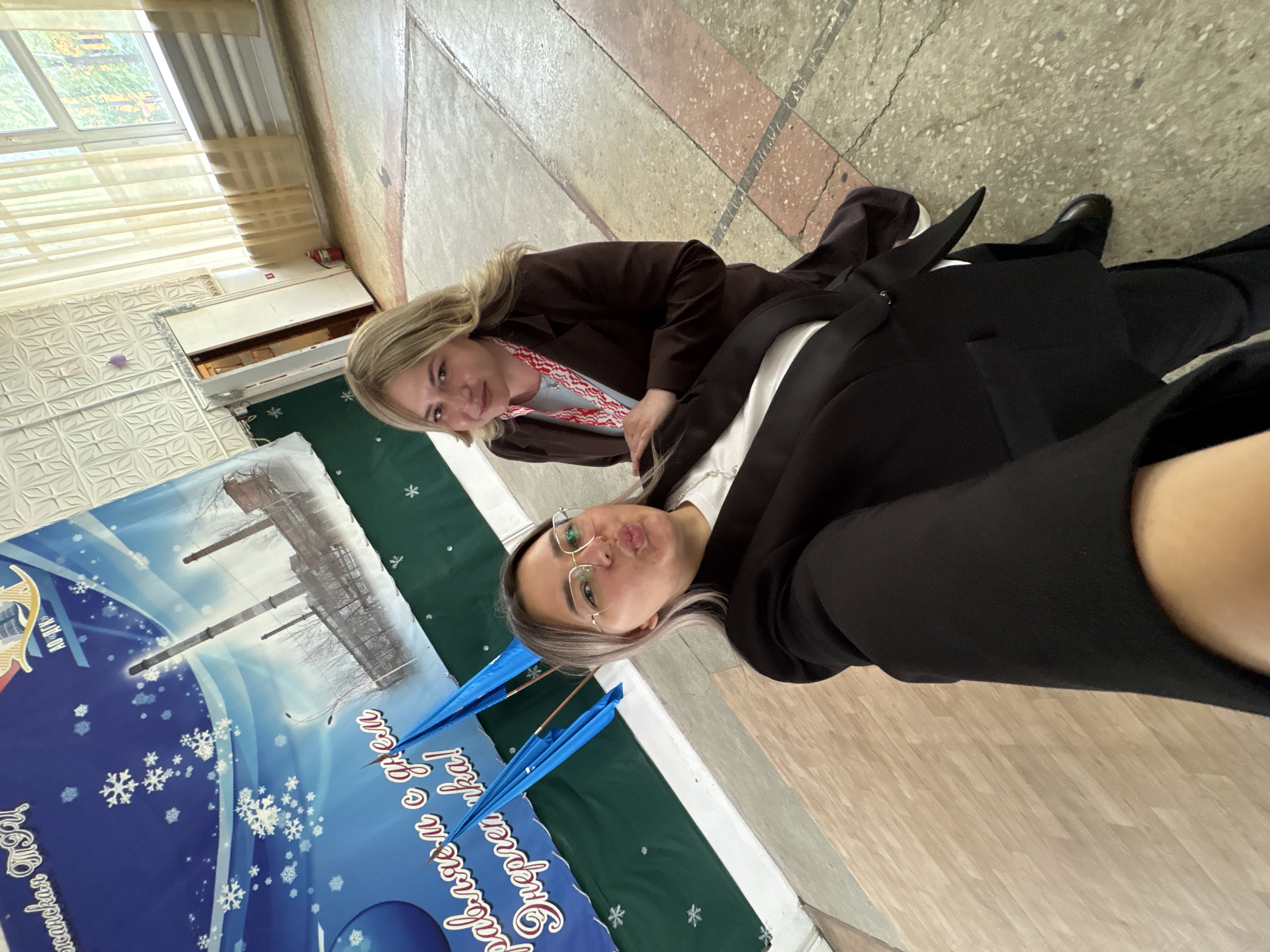 Two women are posing for a selfie. One woman has blonde hair and is wearing a brown jacket. The other woman has brown hair, wears glasses, and is wearing a black jacket. They are standing in what appears to be an interior space with a window in the background. Behind them is a banner with a winter-themed design and Russian text, potentially related to Birobidzhan. The floor has a mottled pattern. Both women are well-dressed and appear to be smiling.