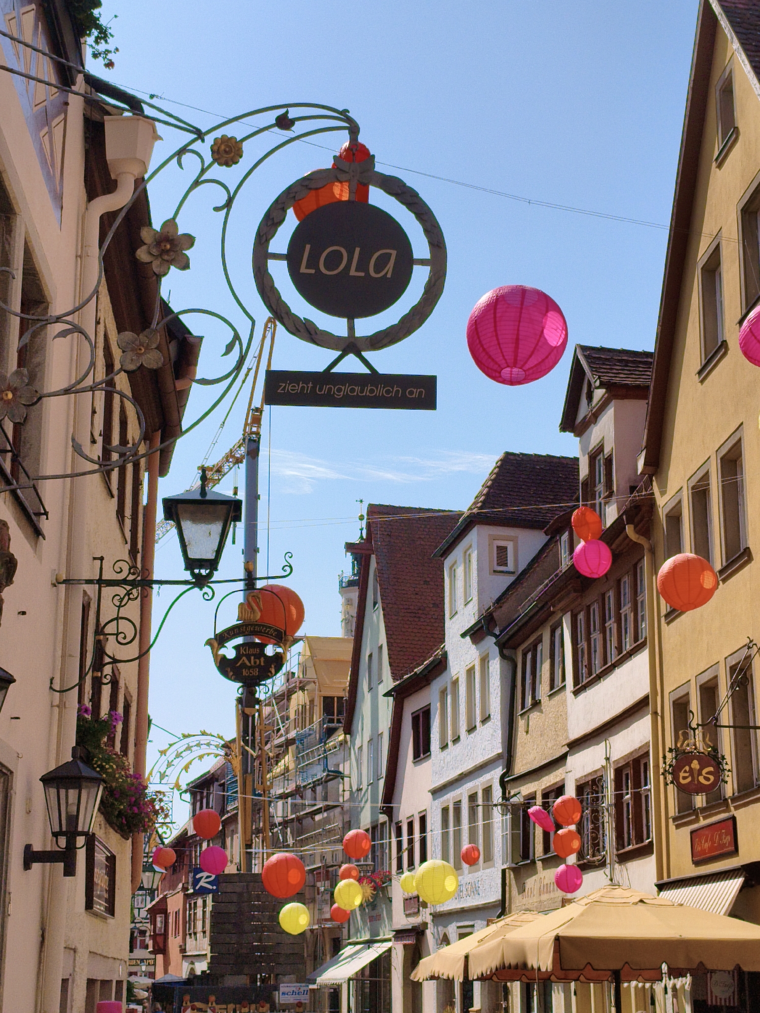 This is a vertical, eye-level shot of a narrow cobblestone street in Rothenburg ob der Tauber, Germany, on a clear, sunny day. The street is lined with traditional half-timbered buildings, some with stucco facades in shades of yellow and white, and others with darker wood beams. Decorative wrought-iron signs hang from many of the buildings. A variety of colorful paper lanterns, primarily in pink and orange, are strung across the street, some illuminated by the sun and others appearing as decorative elements.
In the foreground, to the left, is a building with a cream-colored facade and dark windows. A black wrought-iron lantern hangs from a decorative bracket extending from this building. Further down the street, another building with a darker facade is visible, also adorned with a hanging sign and more lanterns. To the right, a yellow building with multiple windows dominates the right side of the frame. A canvas awning extends from one of the buildings on the right, casting a shadow on the street.
Several signs are visible, including one with the text "LOLA" within a circular frame, and another below it reading "zieht unglaublich an". Another sign displays "Kunstgewerbe Klaus Abt 1658". A sign with "eis" is also visible on the right.
In the background, a yellow construction crane is visible between buildings, suggesting some ongoing work or renovation. The sky is a clear, bright blue with no clouds. The overall impression is a charming, picturesque street scene with vibrant decorations. No people are clearly visible in the image.