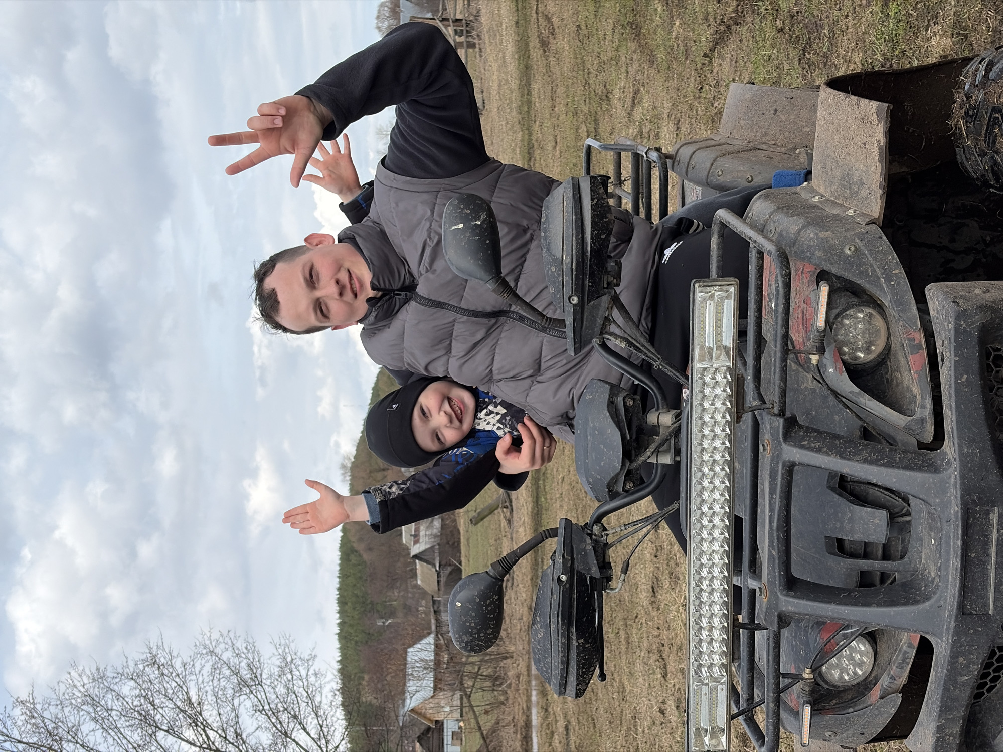 Two individuals, an adult and a child, are positioned on an all-terrain vehicle (ATV) amidst a rural landscape. The adult is seated behind and making a "peace" sign with their hand. The child is positioned in front of the adult and extending their arm outwards. The ATV is heavily soiled with mud, obscuring details of its make and model. The surrounding environment consists of a grassy field with sparse trees. Buildings are visible in the background, suggesting the location might be near or within a small village or rural settlement. The overcast sky and bare branches of the trees suggest it is either late autumn or early spring. The location is Кемуль, Russia.