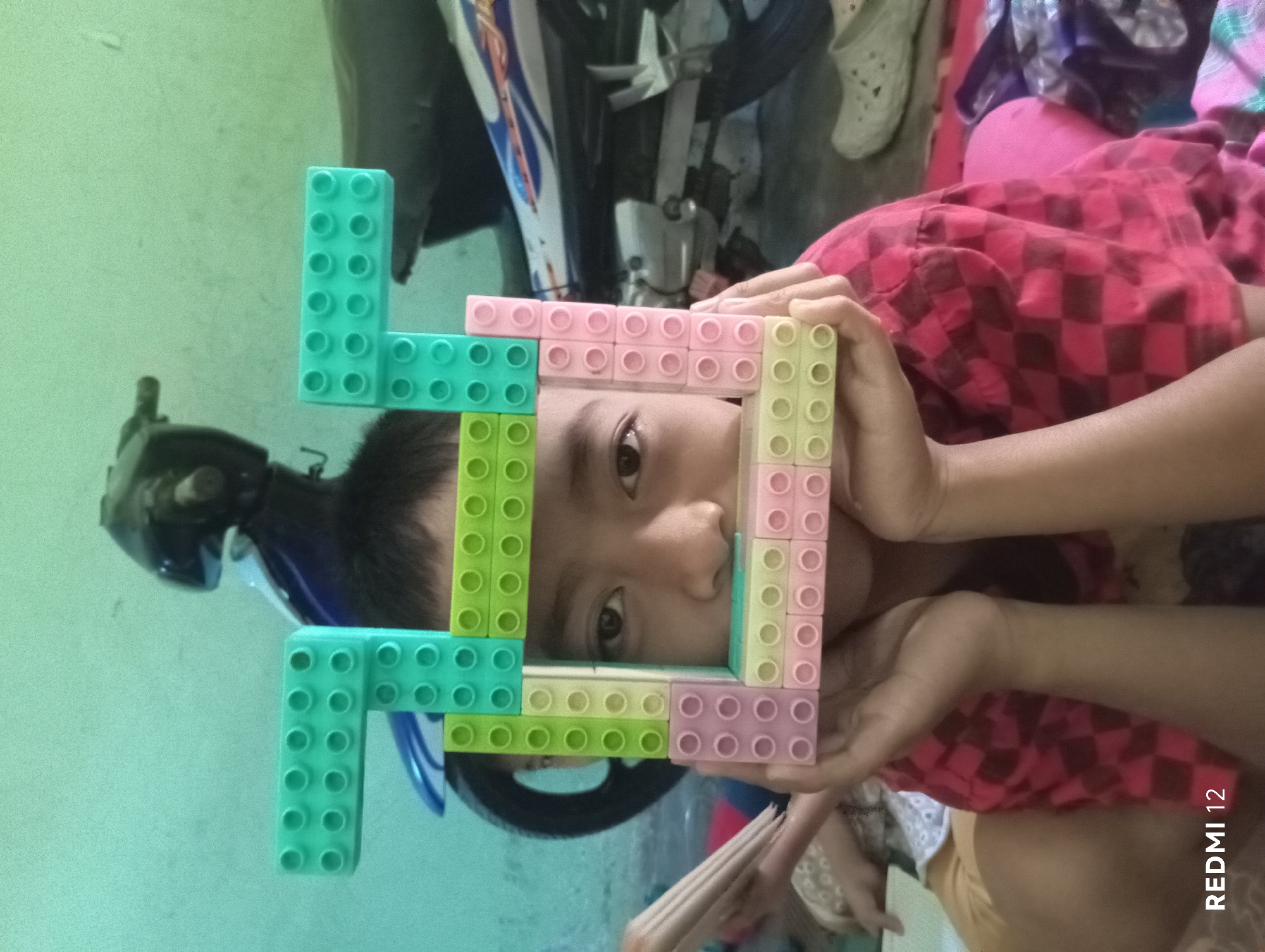 A young person is shown from the chest up, holding a frame constructed from colorful building blocks in front of their face, obscuring their mouth and nose. The blocks are predominantly pastel shades of pink, yellow, and light green, with some teal blocks forming the top of the frame. The individual has dark hair and appears to be looking directly at the camera through the opening in the block frame. They are wearing a red shirt with a dark checkered pattern. The background is indistinct, but a motorcycle is visible behind the person. The overall lighting suggests an indoor setting. The image is watermarked "REDMI 12" in the lower left corner. The scene is likely a casual, personal moment captured within a residence or public space in Pagar Alam, Indonesia, given the "REDMI 12" watermark, which is a common mobile phone brand in the region.